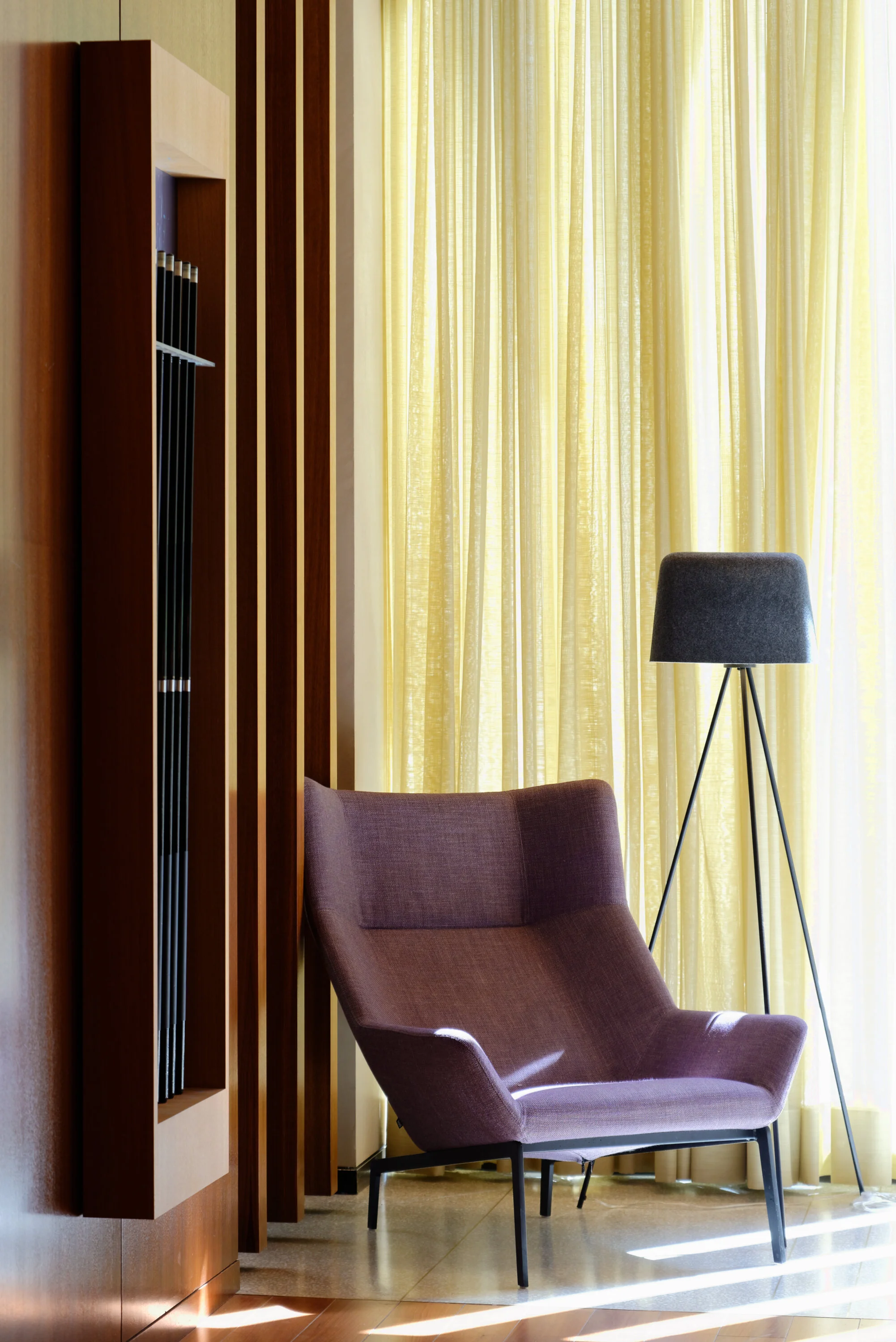 JGNA residential lounge detail with a lilac armchair, tripod floor lamp, and sheer curtains against walnut paneling