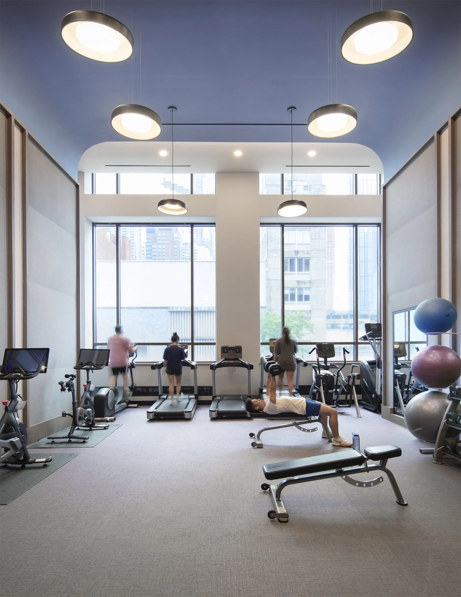 JGNA residential fitness center with cardio equipment and a weight bench beside double-height windows