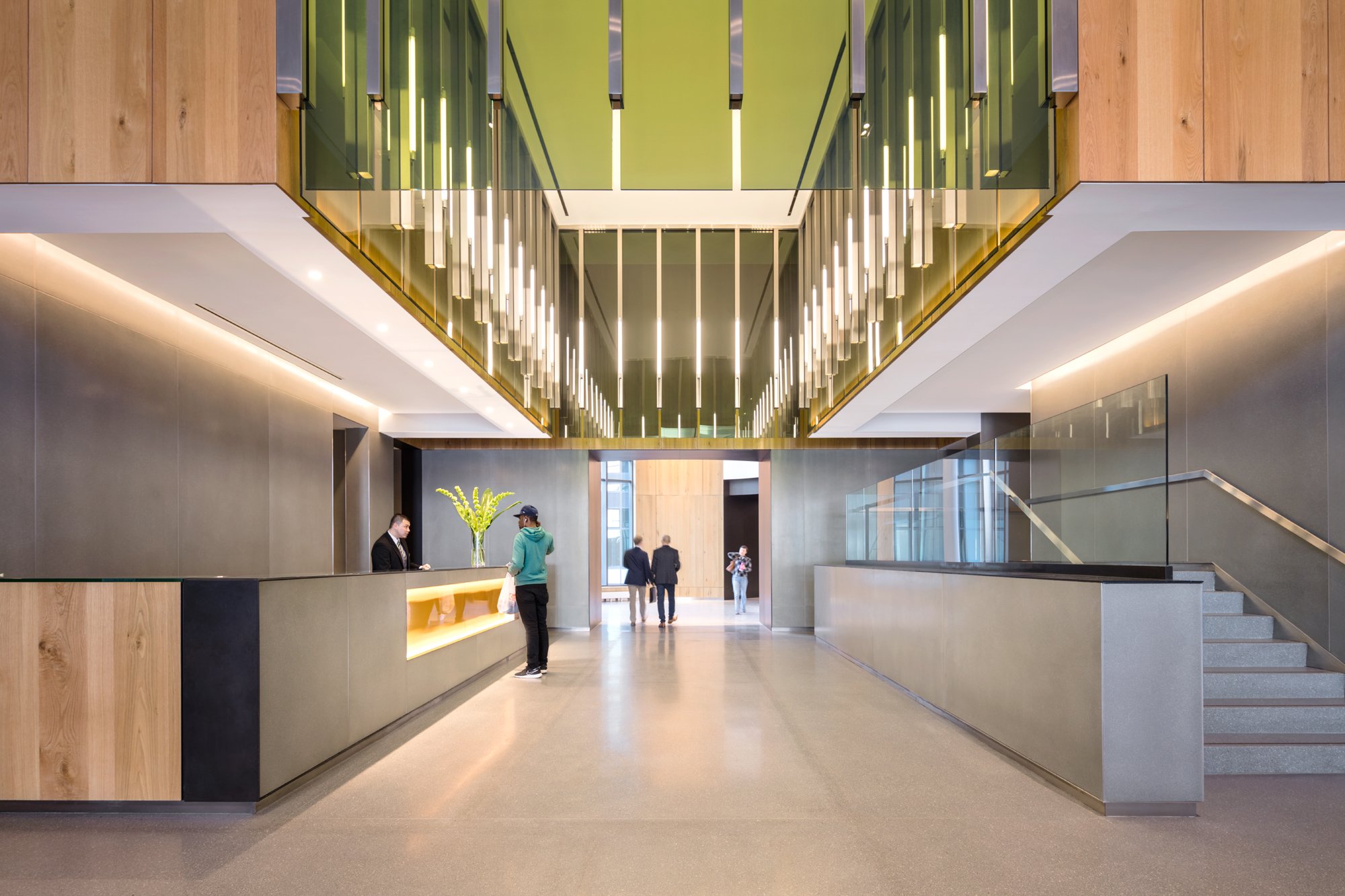 JGNA residential lobby reception desk beneath a double-height brass-framed pendant installation with a stair beyond