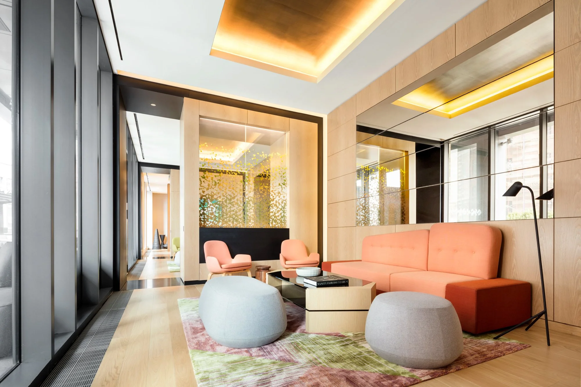 JGNA residential lounge with a coral sofa, pink armchairs, felt poufs, and a patterned rug beside a long corridor