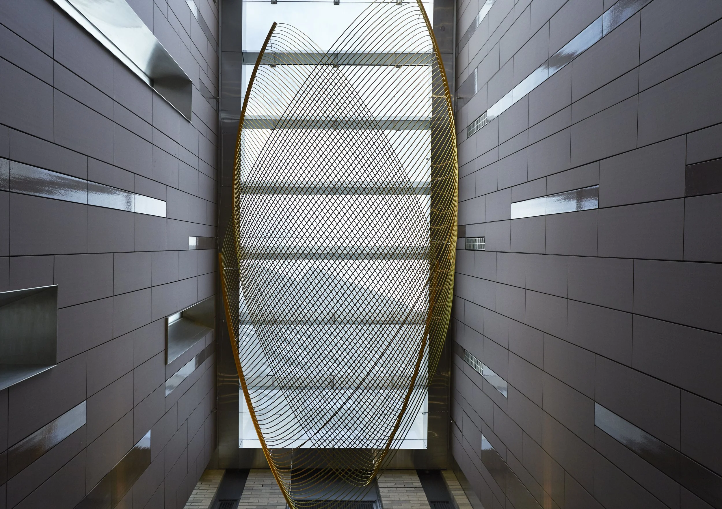 JGNA residential atrium with grey tile-clad walls and a suspended golden mesh sculpture in a skylit shaft