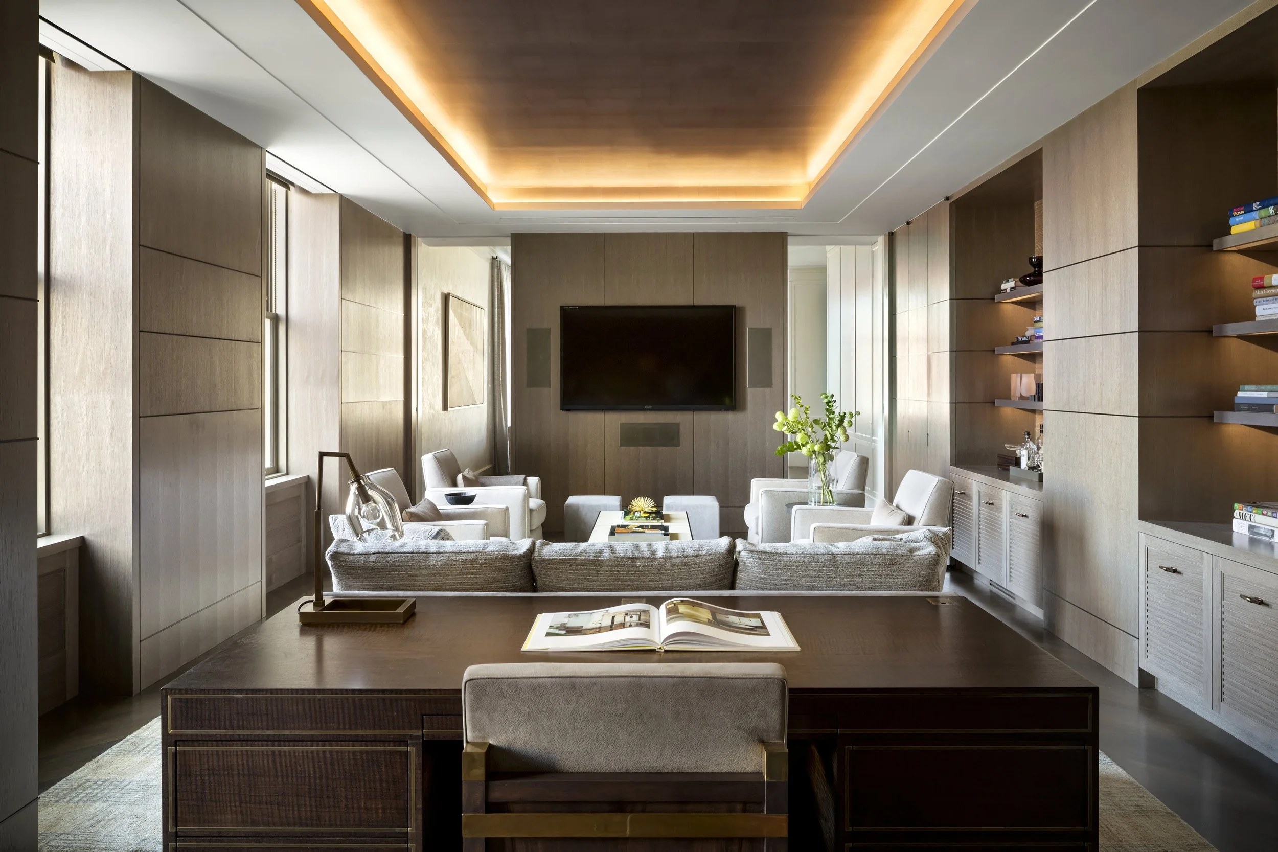JGNA residence study with a dark wood desk, neutral lounge seating, backlit shelving, and a cove-lit ceiling
