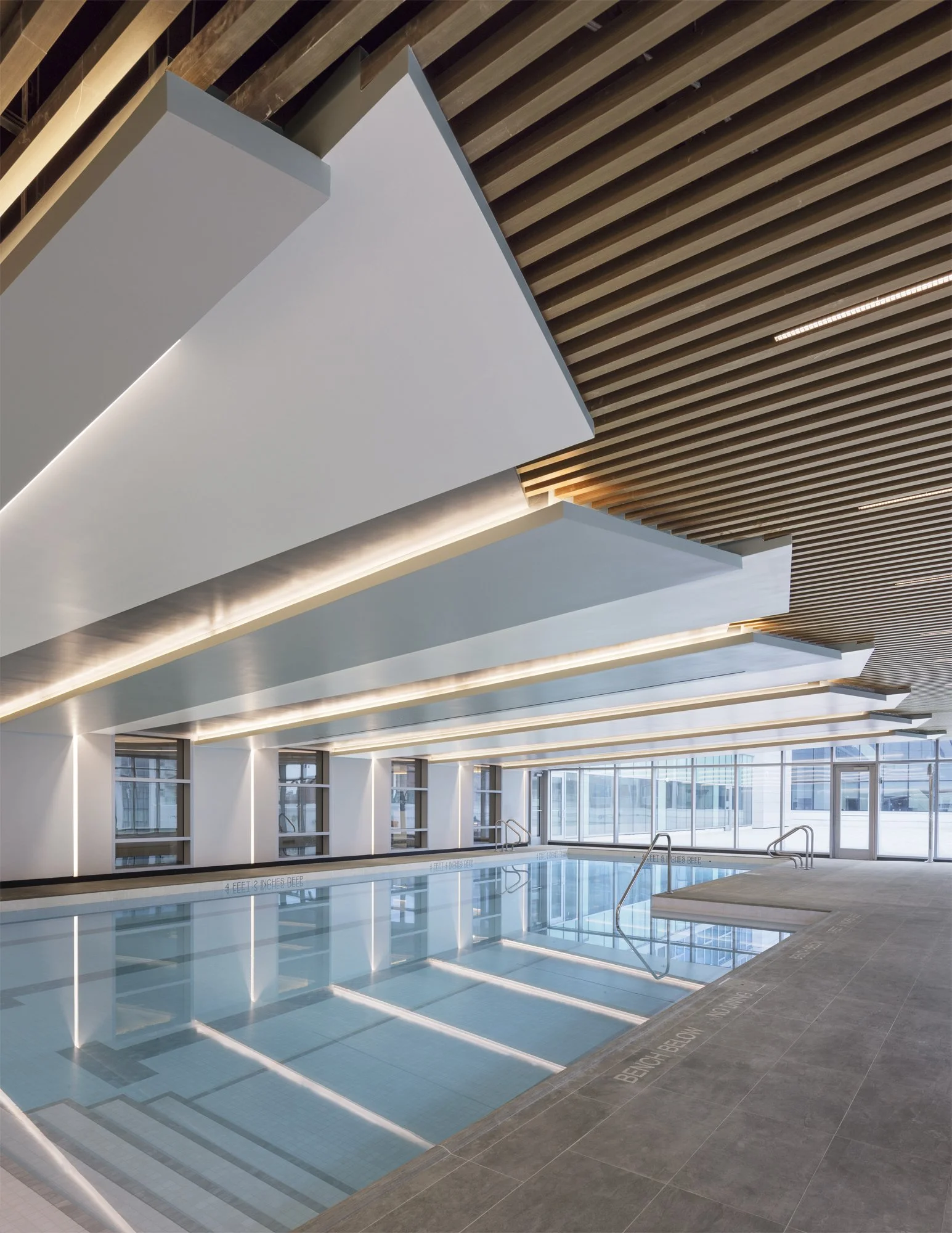 JGNA residential indoor pool with angular white soffits, a wood-slat ceiling, and cove lighting over the water