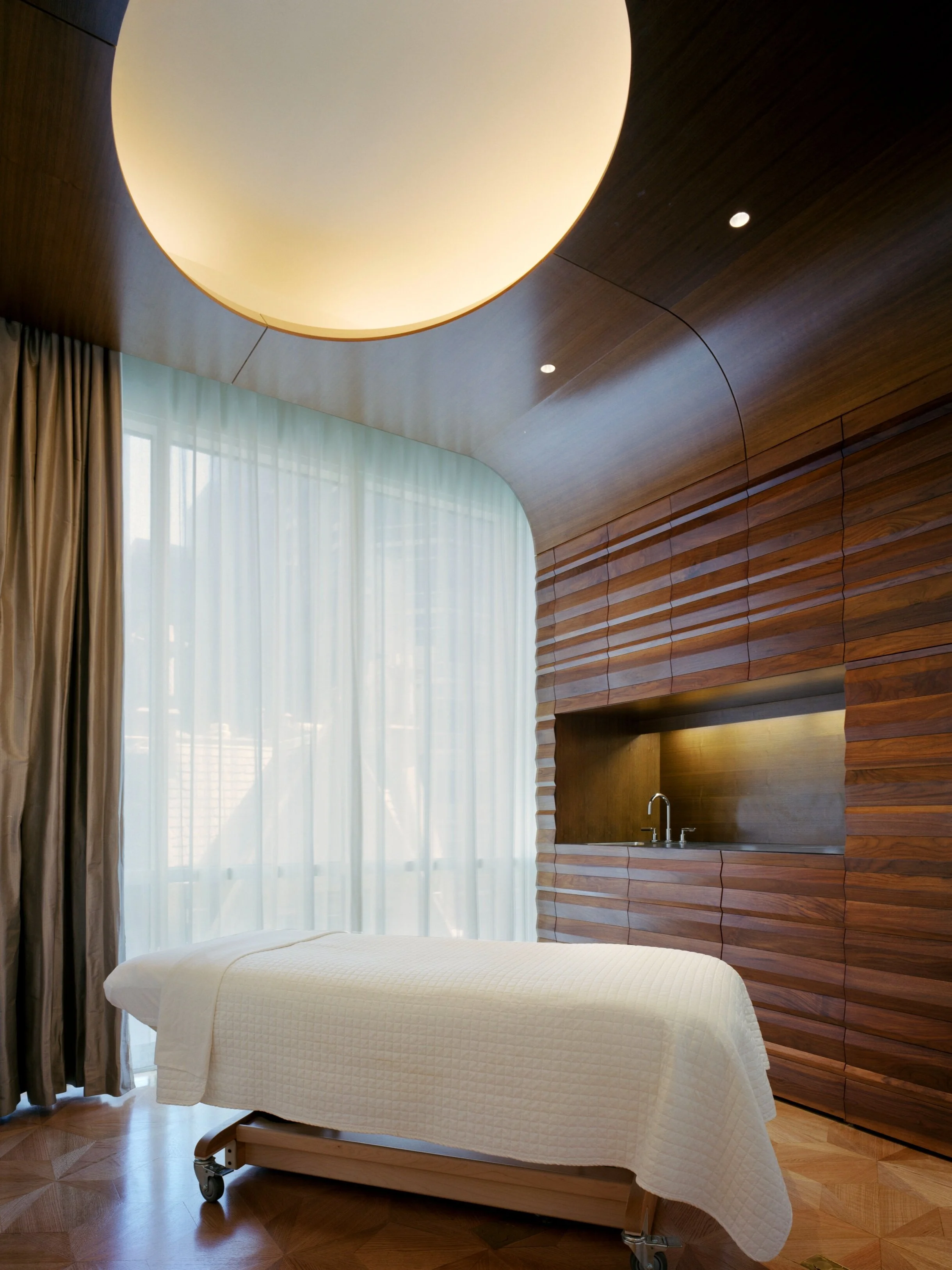 JGNA spa treatment room with walnut paneling, a backlit ceiling disc, sheer curtains, and a massage table