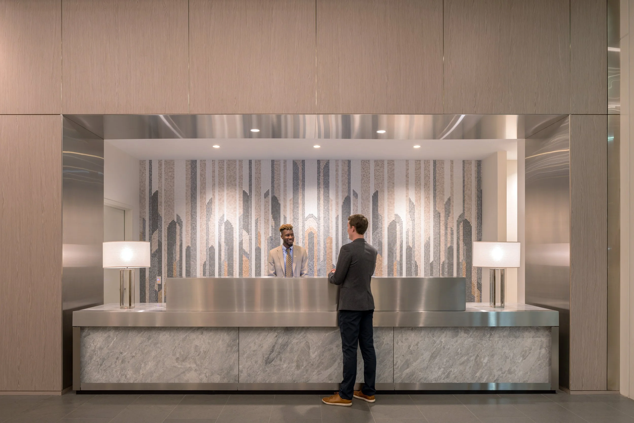 JGNA residential lobby reception desk with a stainless surround, marble base, and a city-skyline glass mosaic mural