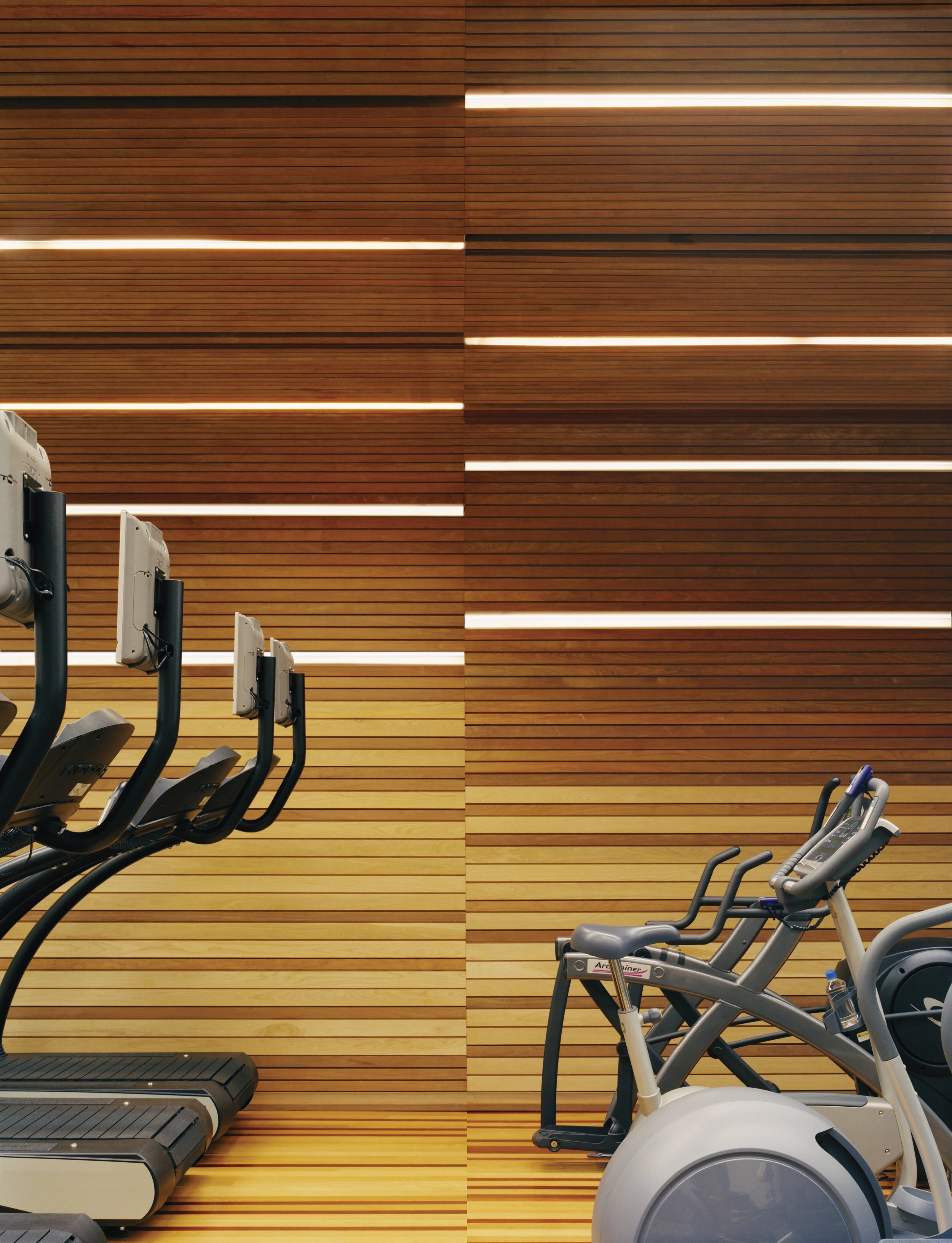 JGNA fitness center with cardio machines against a backlit horizontal wood-slat wall