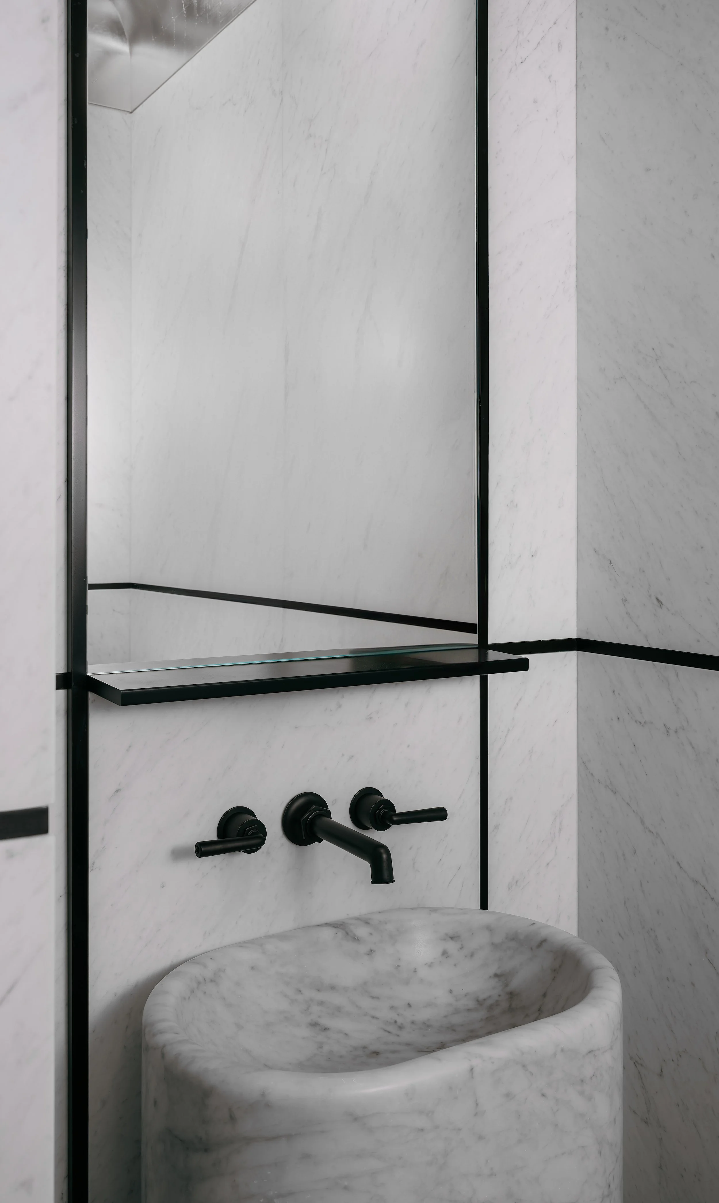 Powder room clad in white marble with a cylindrical pedestal sink and black wall-mounted faucet
