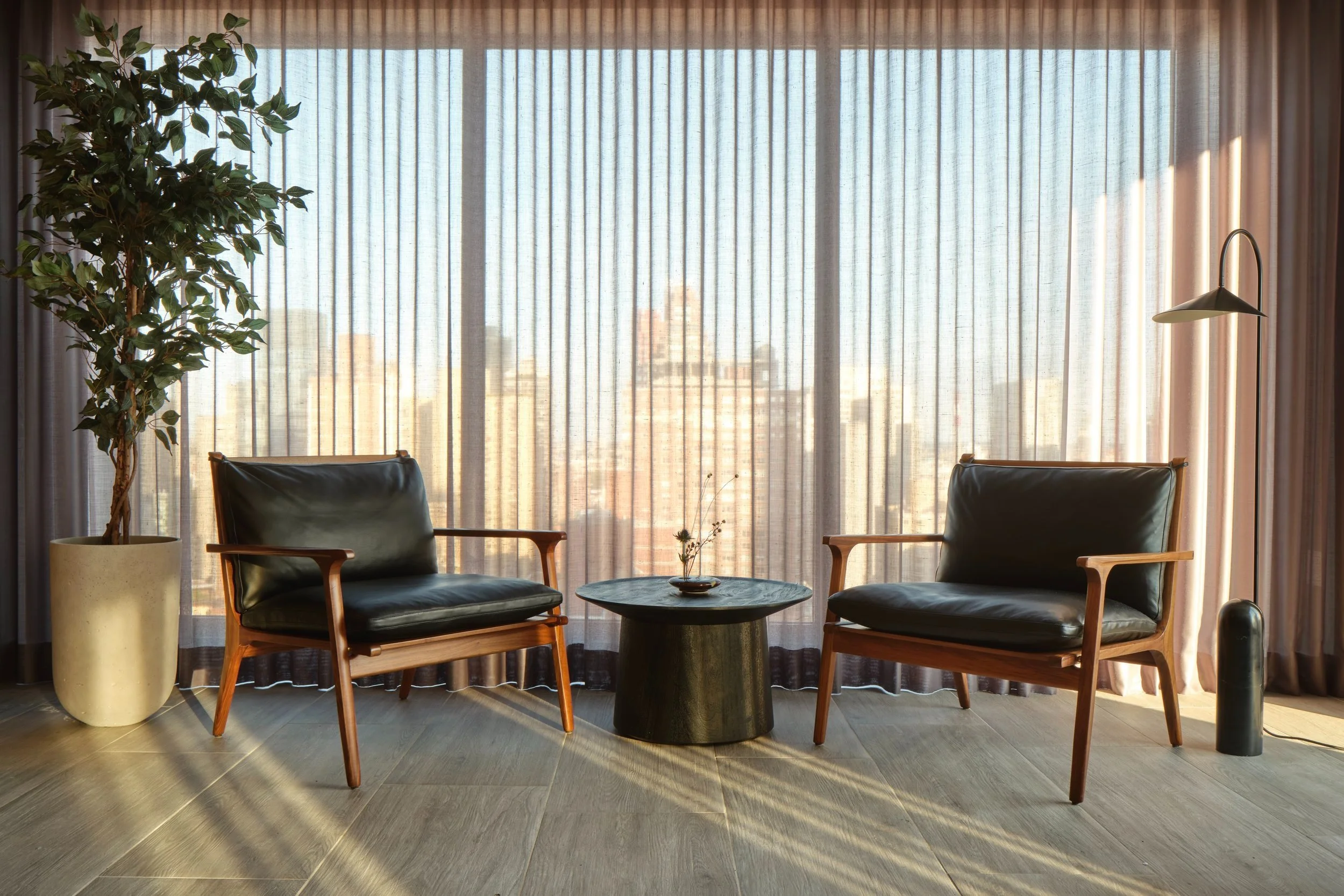JGNA residential lounge with paired black leather armchairs, a bronze drum table, and sheer-curtained windows