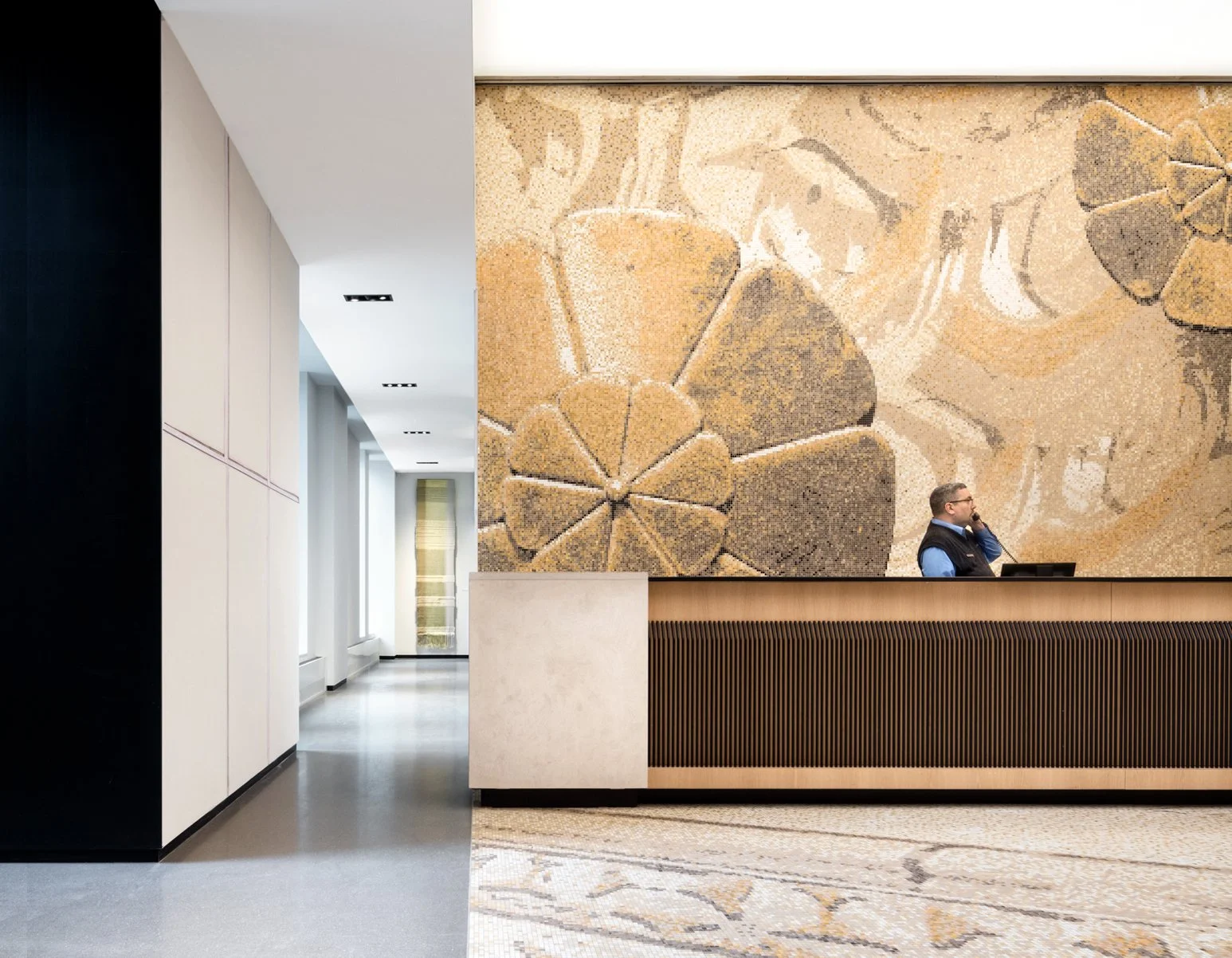 JGNA residential lobby reception desk with a ribbed wood front, a botanical mosaic mural, and a terrazzo floor