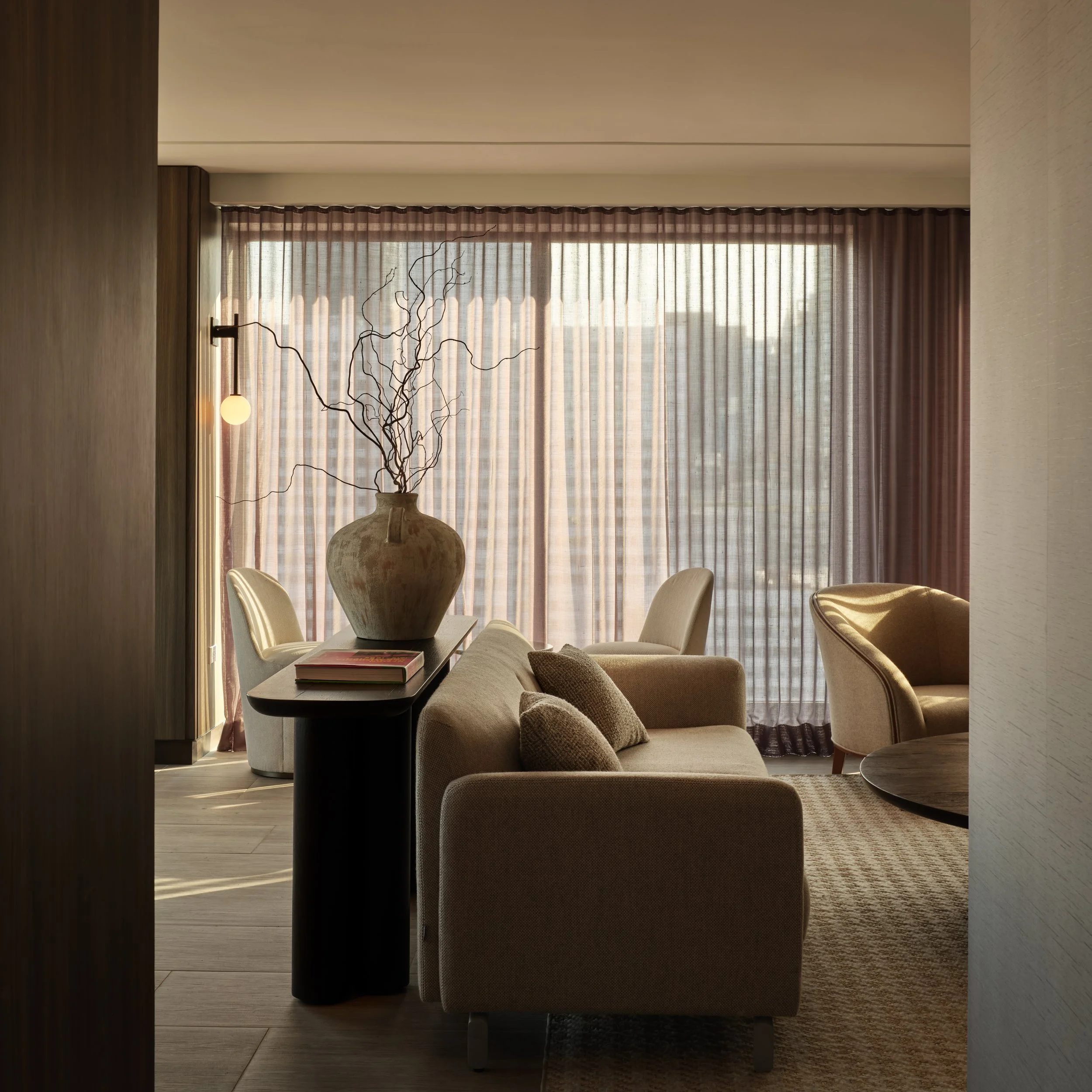 JGNA residential lounge with bouclé seating, a sculptural console, sheer curtains, and a warm city view at sunset