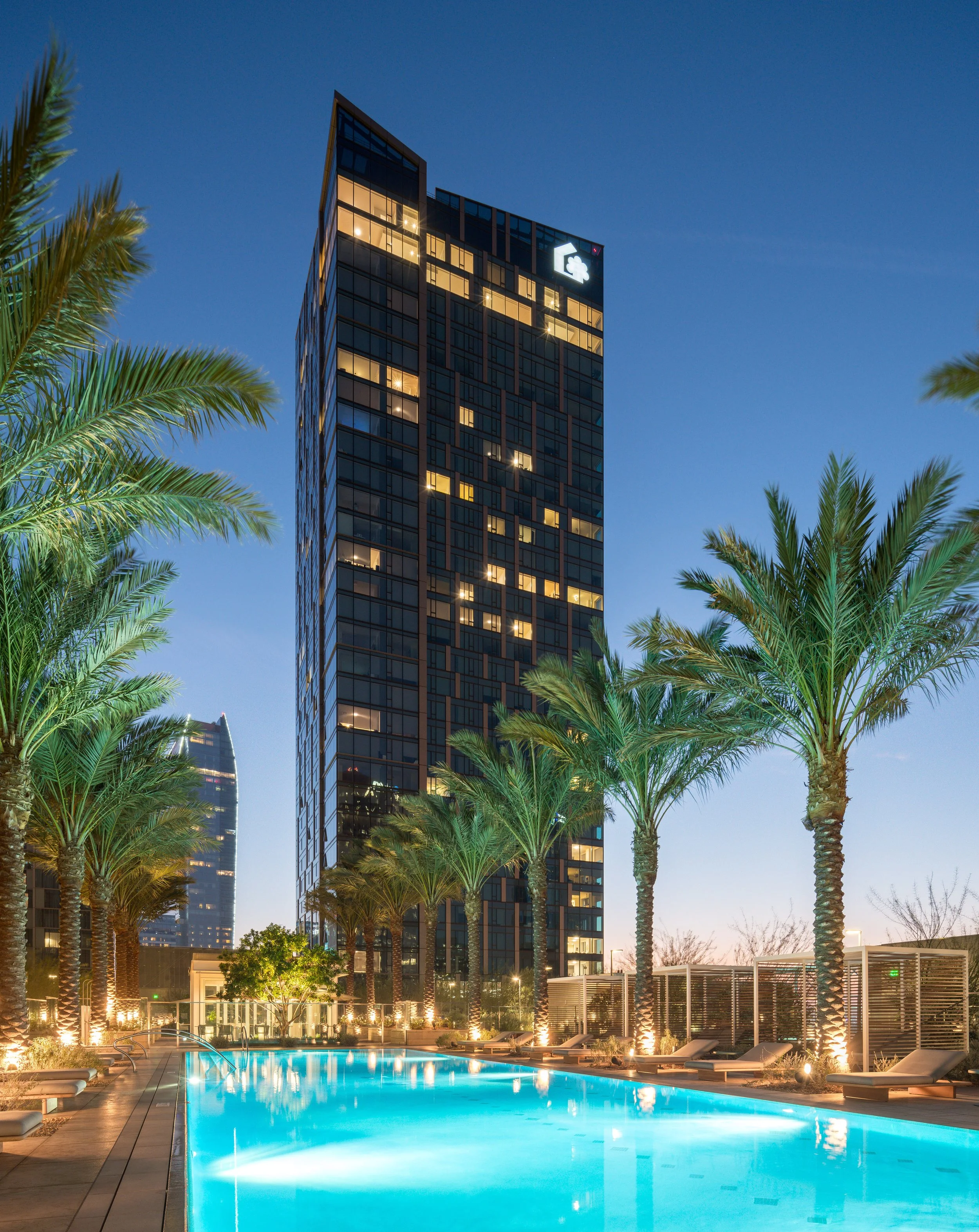 JGNA residential tower exterior at dusk with a lit infinity pool, loungers, and palm trees on the deck