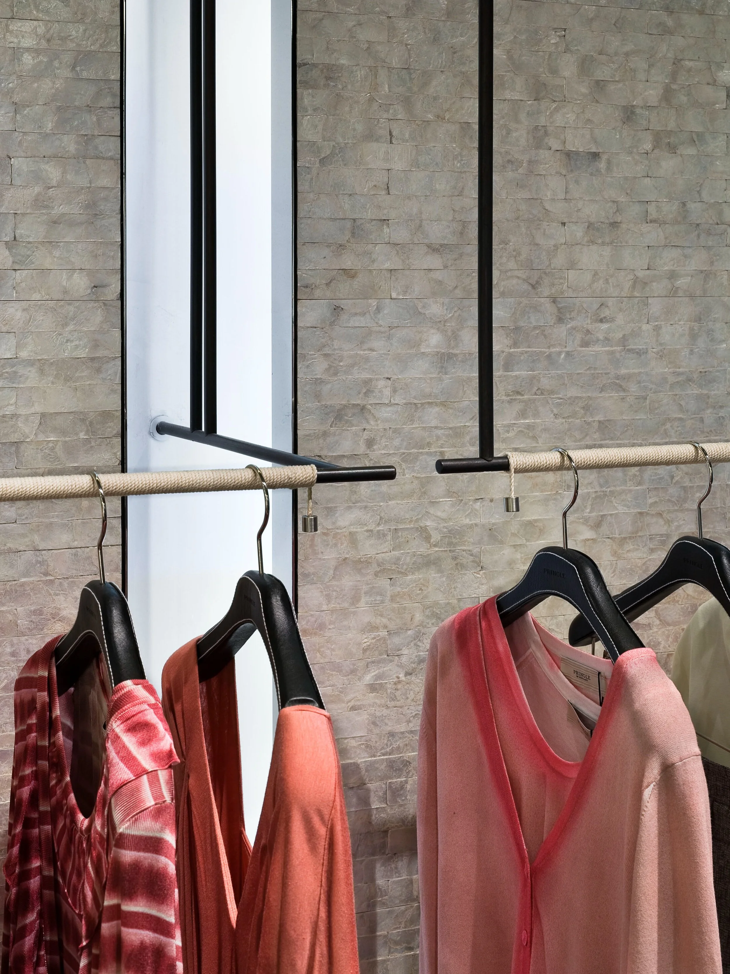 JGNA boutique garment rail of coral and pink clothing against a textured stone wall beside a window