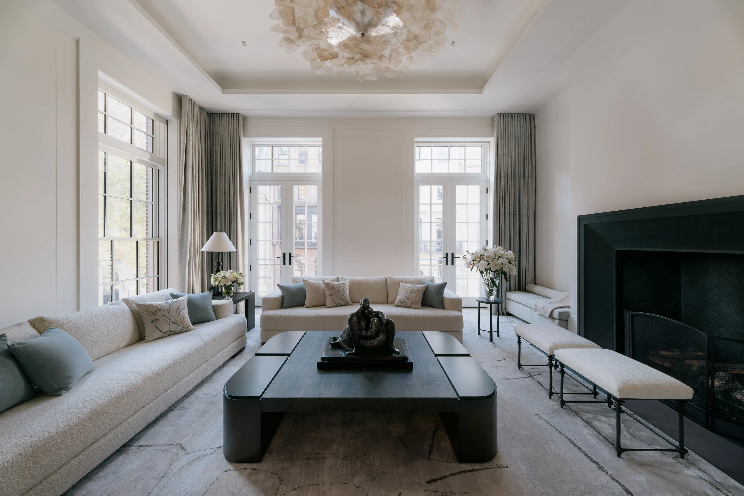 Spacious, light-filled luxury living room featuring tall windows, a striking contemporary glass chandelier, neutral seating, and a dark fireplace in a bespoke Manhattan townhouse