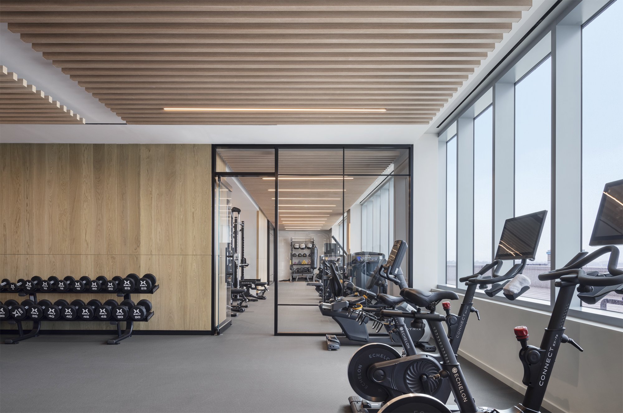 JGNA residential fitness center with free weights, a wood-slat ceiling, spin bikes, and a wall of windows