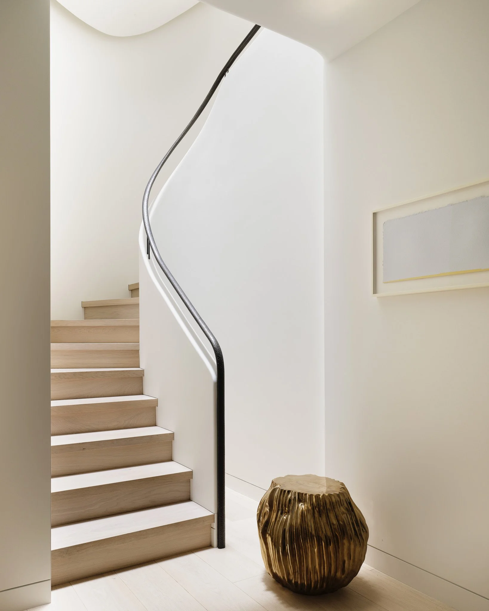 Oak staircase with a curved black handrail and a brass sculptural stool in a West Village townhouse by JG Neukomm Architecture