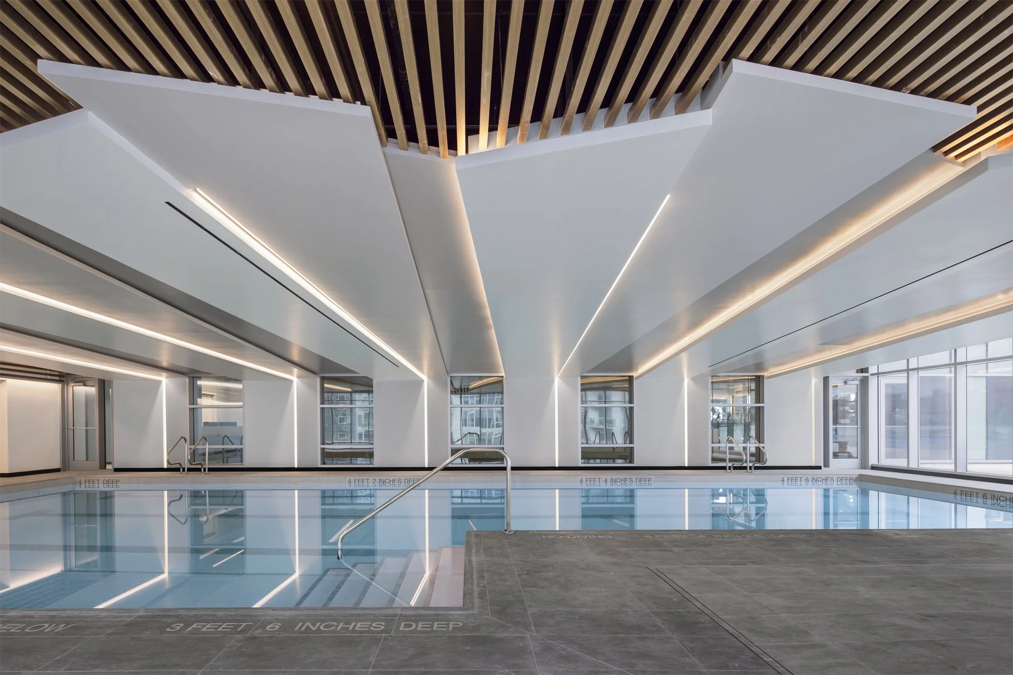 JGNA residential indoor lap pool with recessed linear ceiling lighting, stone decking, and a wall of city windows