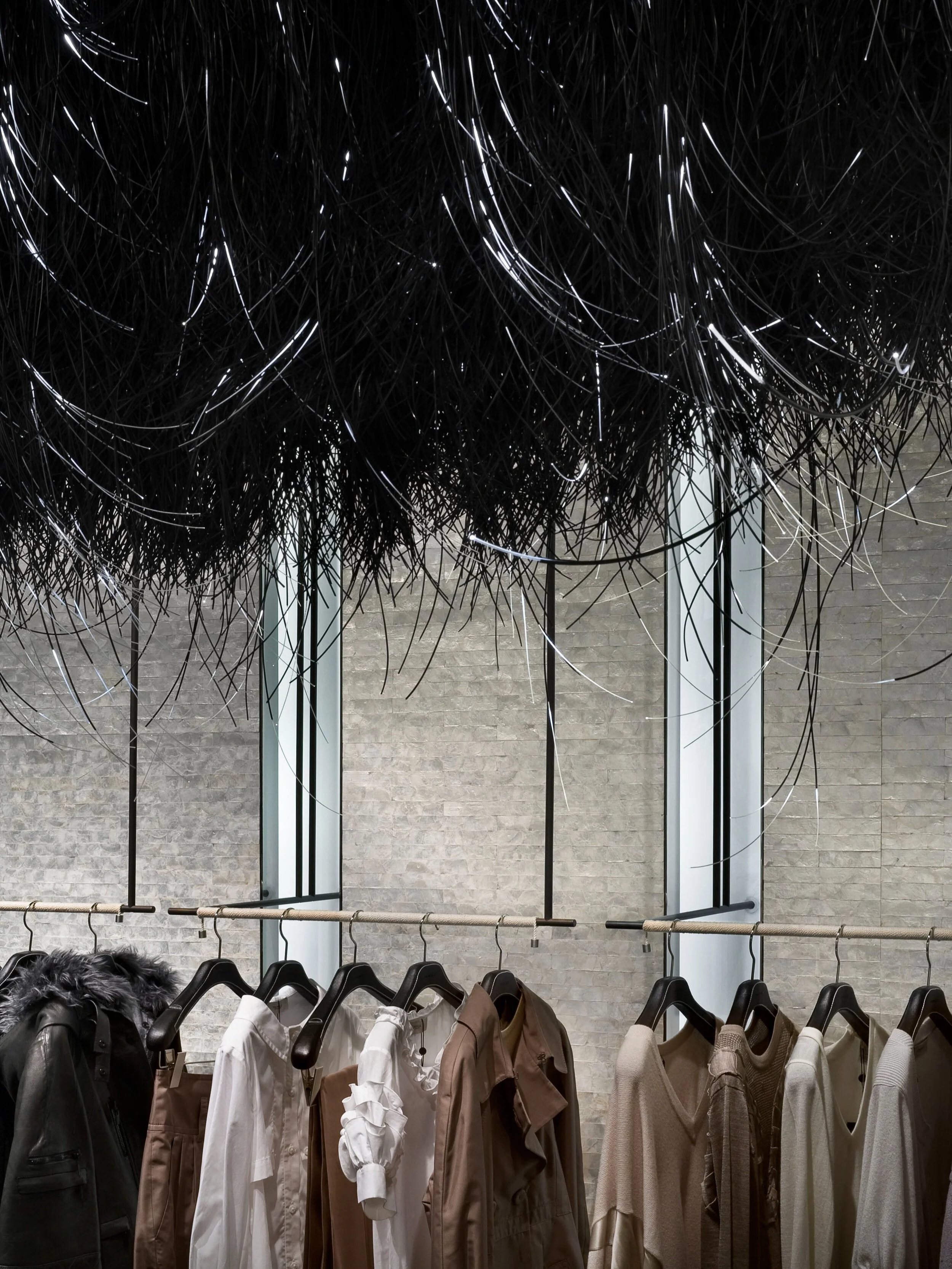 JGNA store interior with a black fringe ceiling installation above a garment rail of neutral-toned clothing