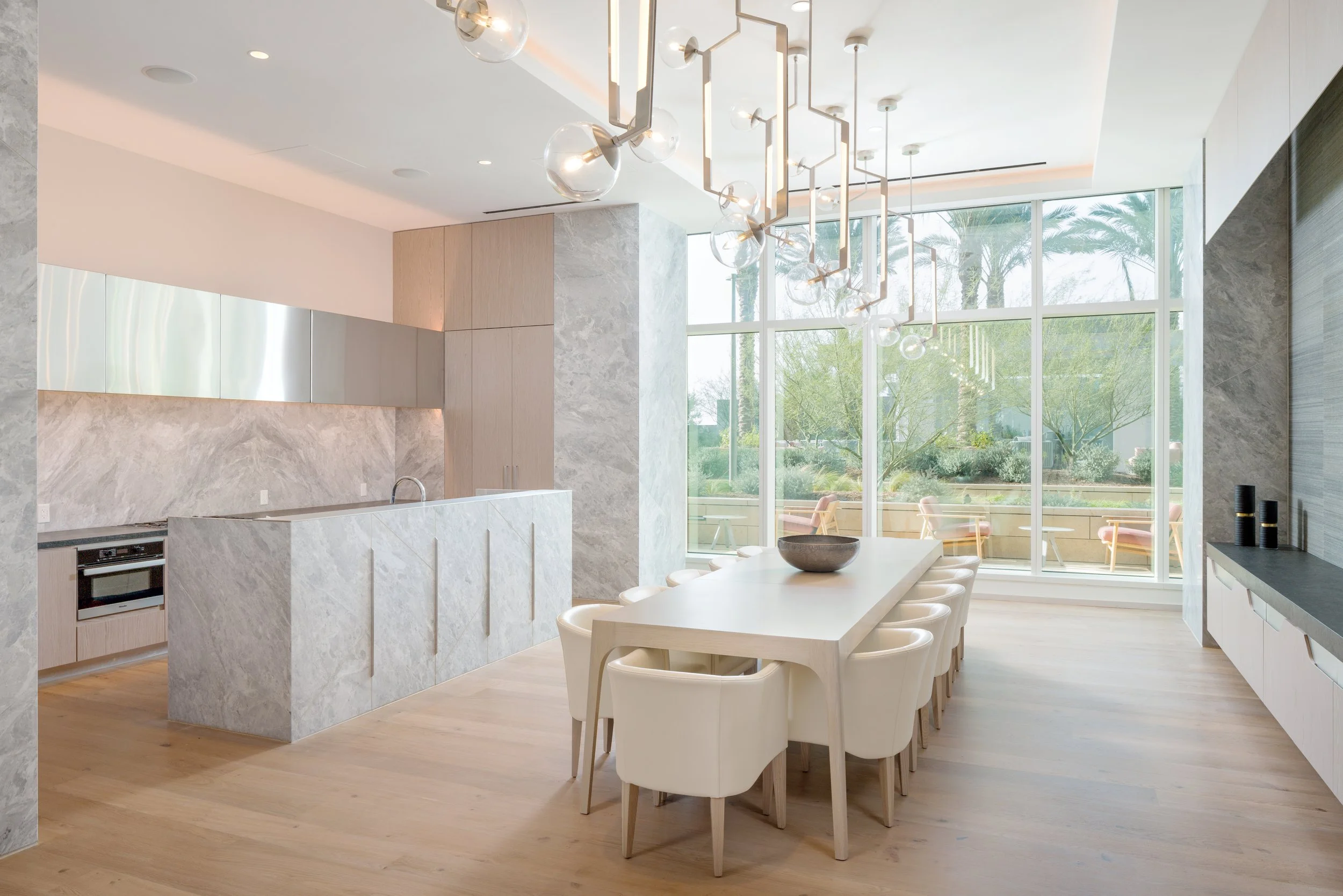 JGNA residential amenity dining room with a marble island, long dining table, globe chandeliers, and floor-to-ceiling windows