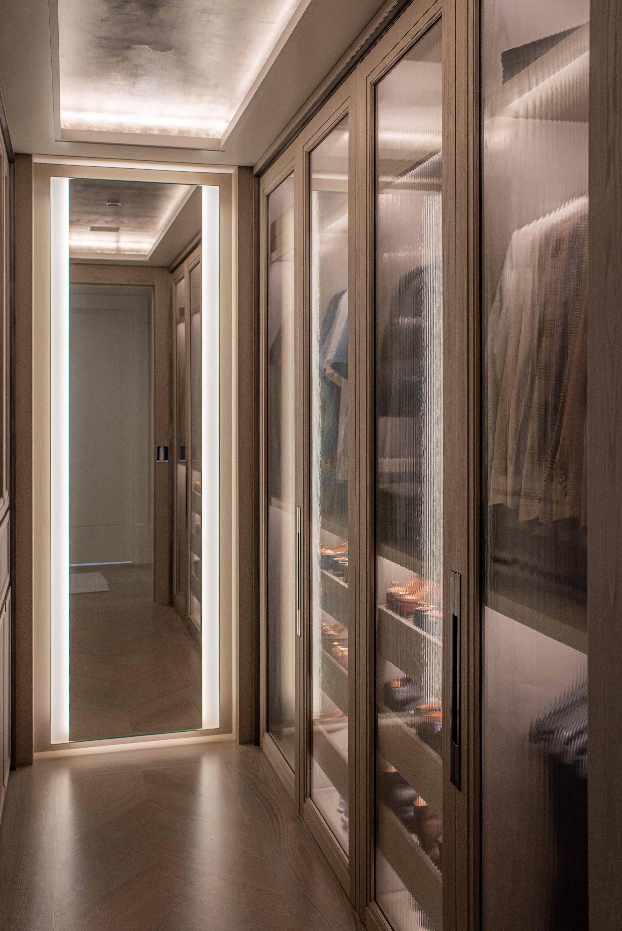 Walk-in dressing room with backlit glass-front wardrobes and herringbone wood flooring