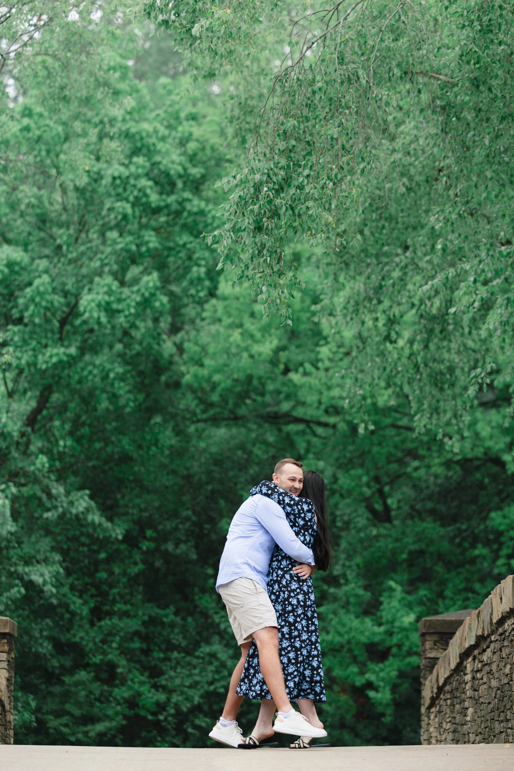 Photo as Boyfriend proposes to his girlfriend on one knee on the duck pond bridge at Freedom Park outdoors in the background after he proposed, by ParksPictures