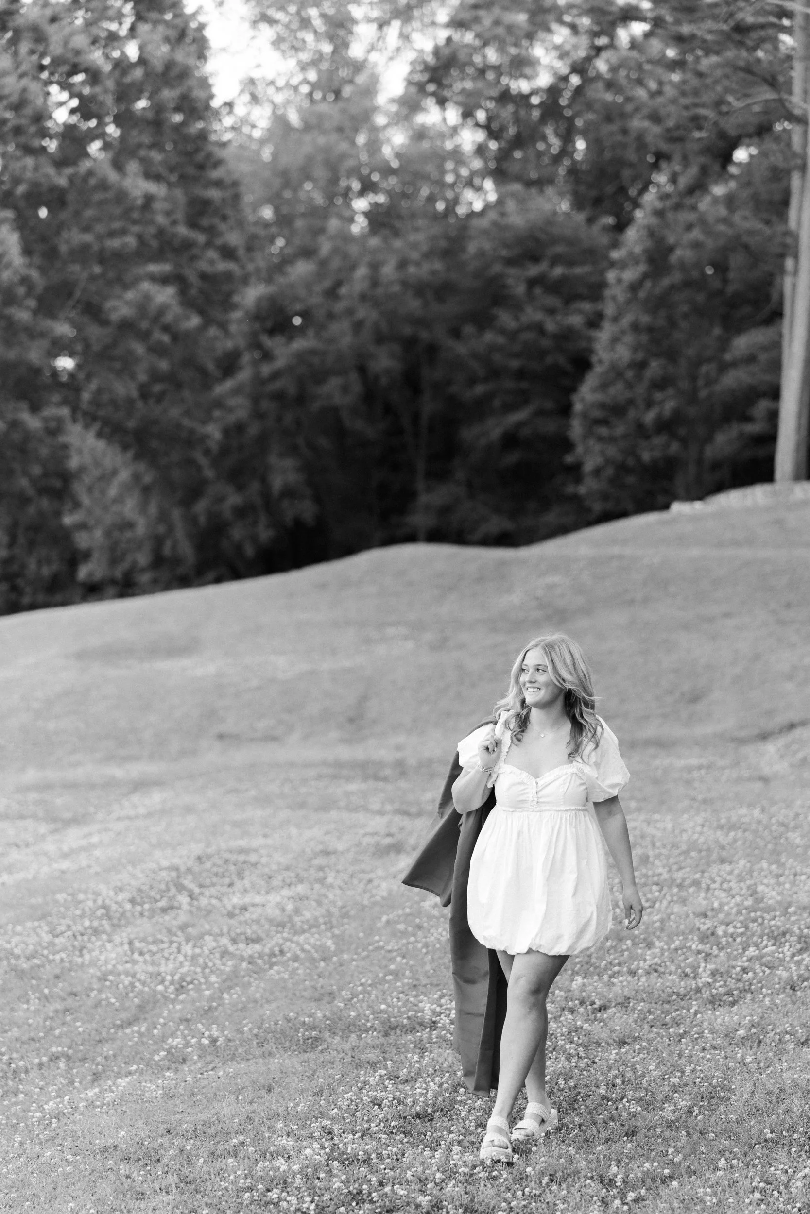 Charlotte Catholic High School Senior girl's senior portraits at Freedom Park in Myers Park Dilworth Charlotte NC in a white ruffle summer dress with sandals walking casual pose with blond hair and graduation gown over shoulder