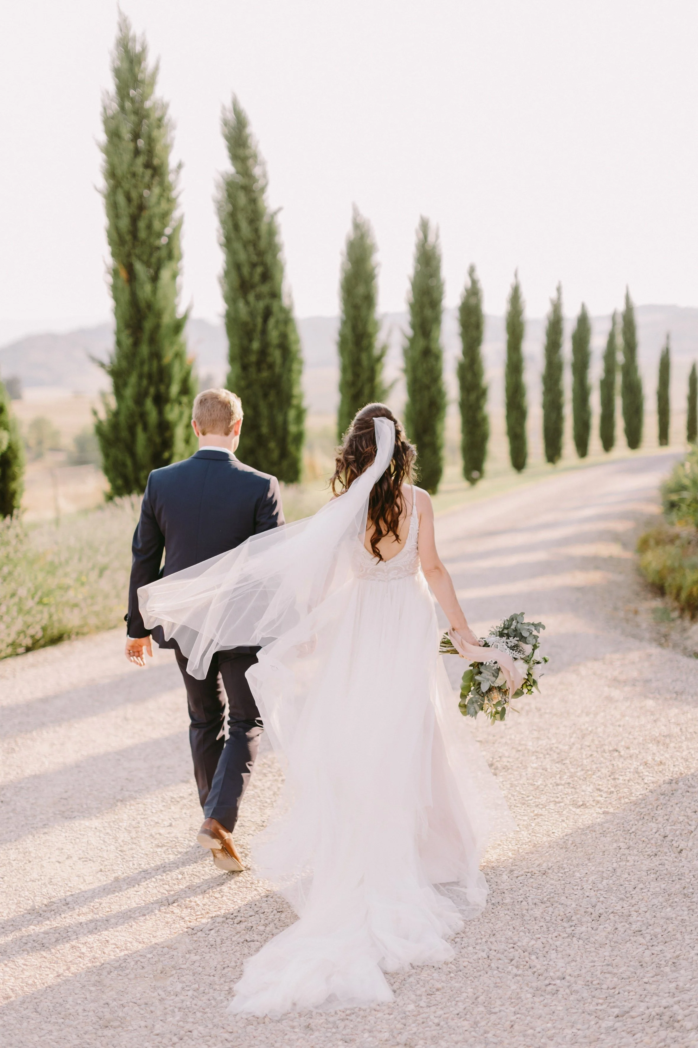 Bride and groom walking on a path lined with tall cypress trees, bride wearing a white gown and holding a bouquet.