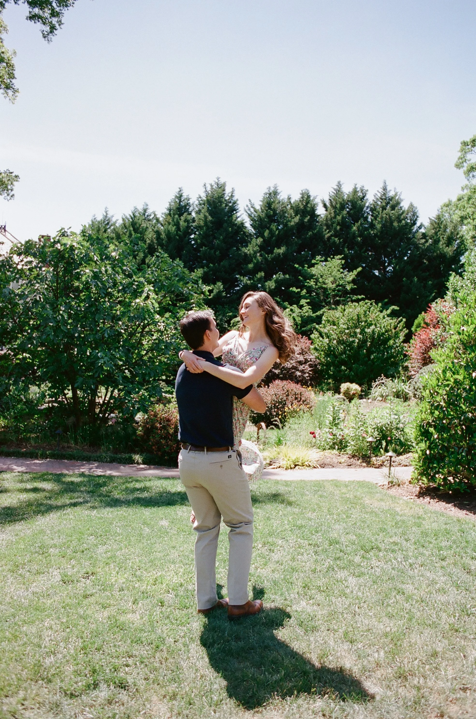 Fiancé swinging his girlfriend around in a summer dress in a field by private gardens at engagement session photographed on Kodak Portra 400 film at Alexander Homestead by Charlotte Wedding Photographer Michael Parks, ParksPictures