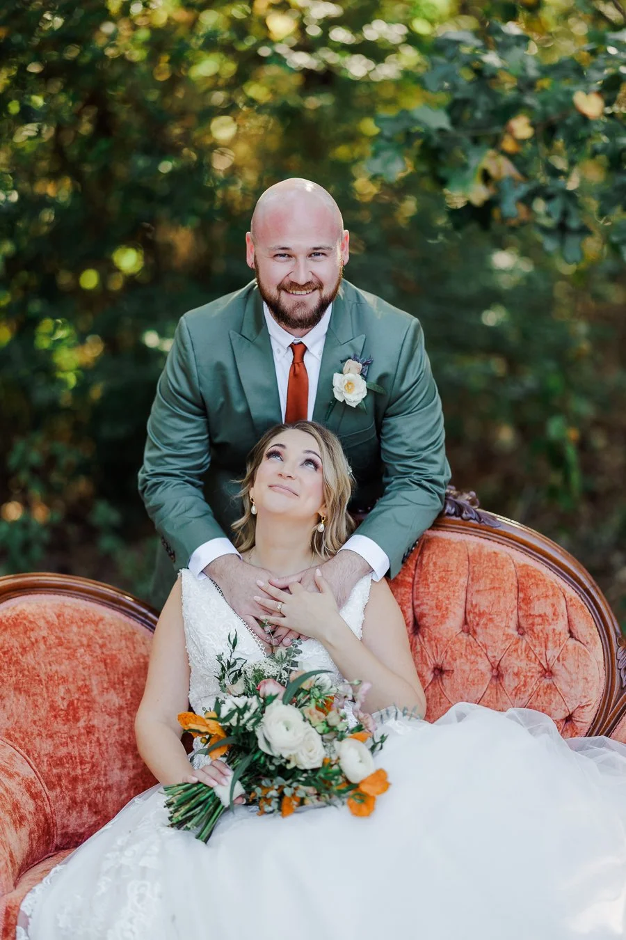 Groom standing over bride and smiling as she looks up from antique overstuffed red couch at their fall Forest Wedding at Il Bella Gardens in Stanley NC by Charlotte Wedding Photographer Michael Parks of ParksPictures