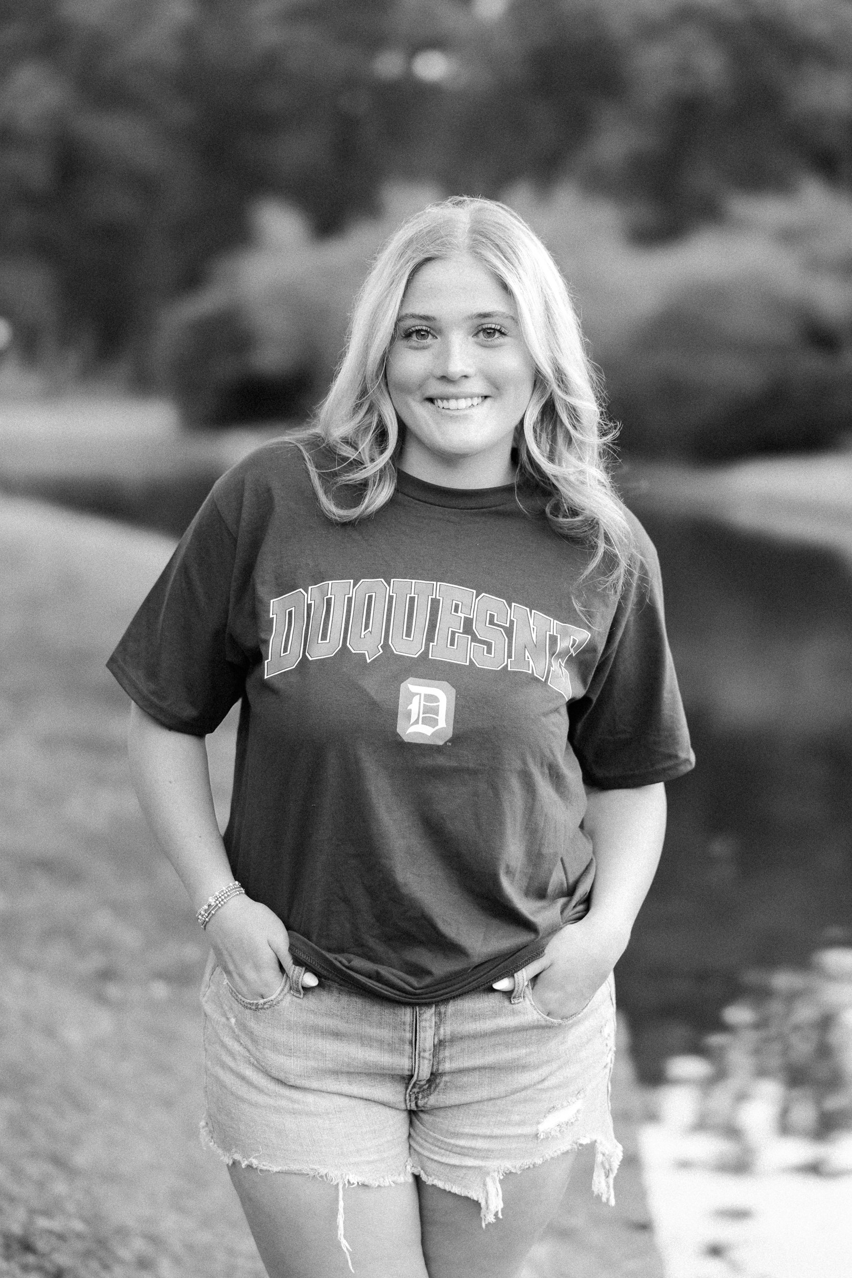 Charlotte Catholic High School Senior girl's senior portraits at Freedom Park in Myers Park Dilworth Charlotte NC wearing a college t-shirt and jean shorts outfit standing in a casual pose with hands in pockets in black and white