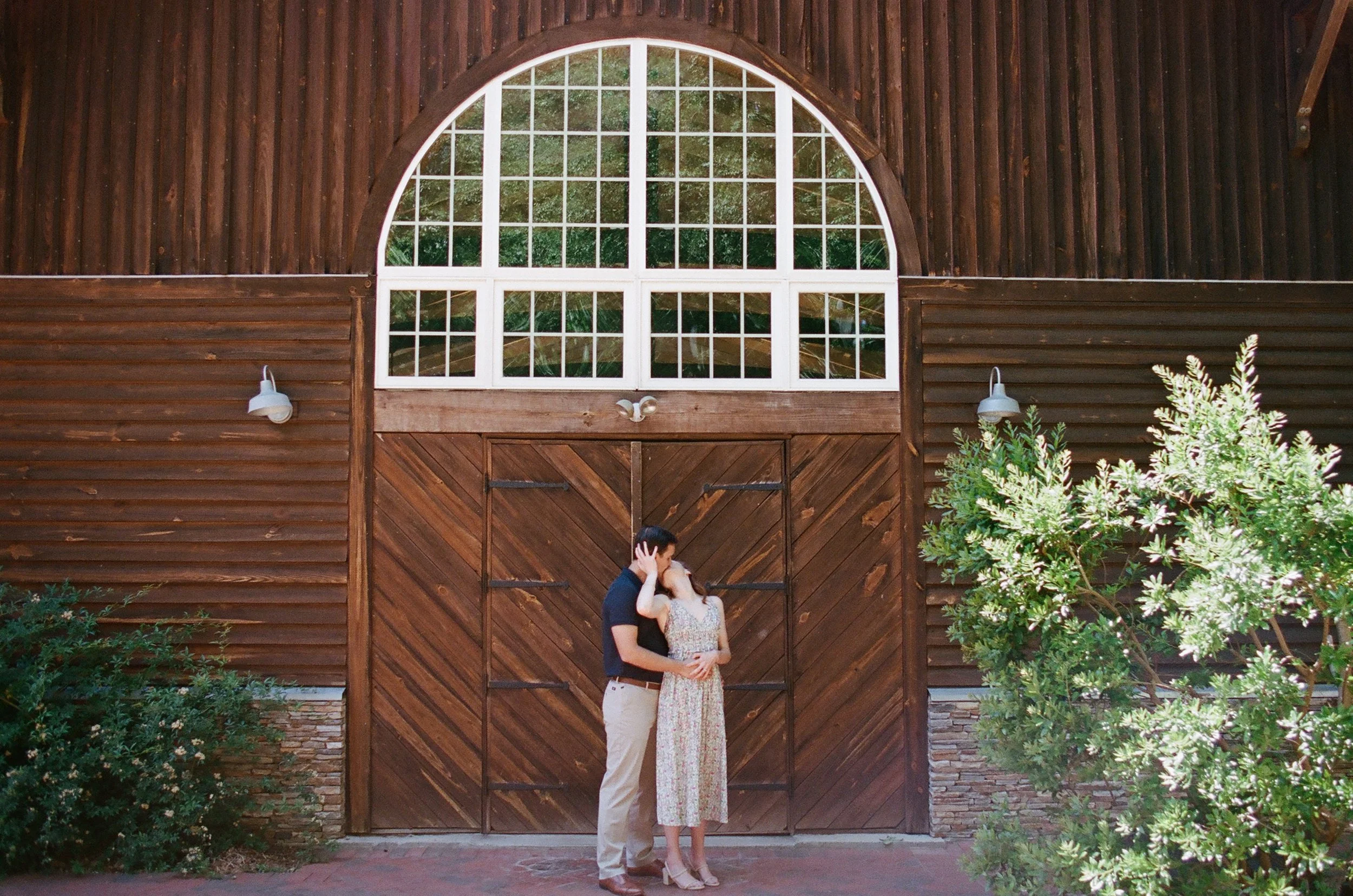 Engaged couple kissing in front of giant barn doors and transom white windows Engagement session photographed on Kodak Portra 400 film at Alexander Homestead by Charlotte Wedding Photographer Michael Parks, owner of ParksPictures