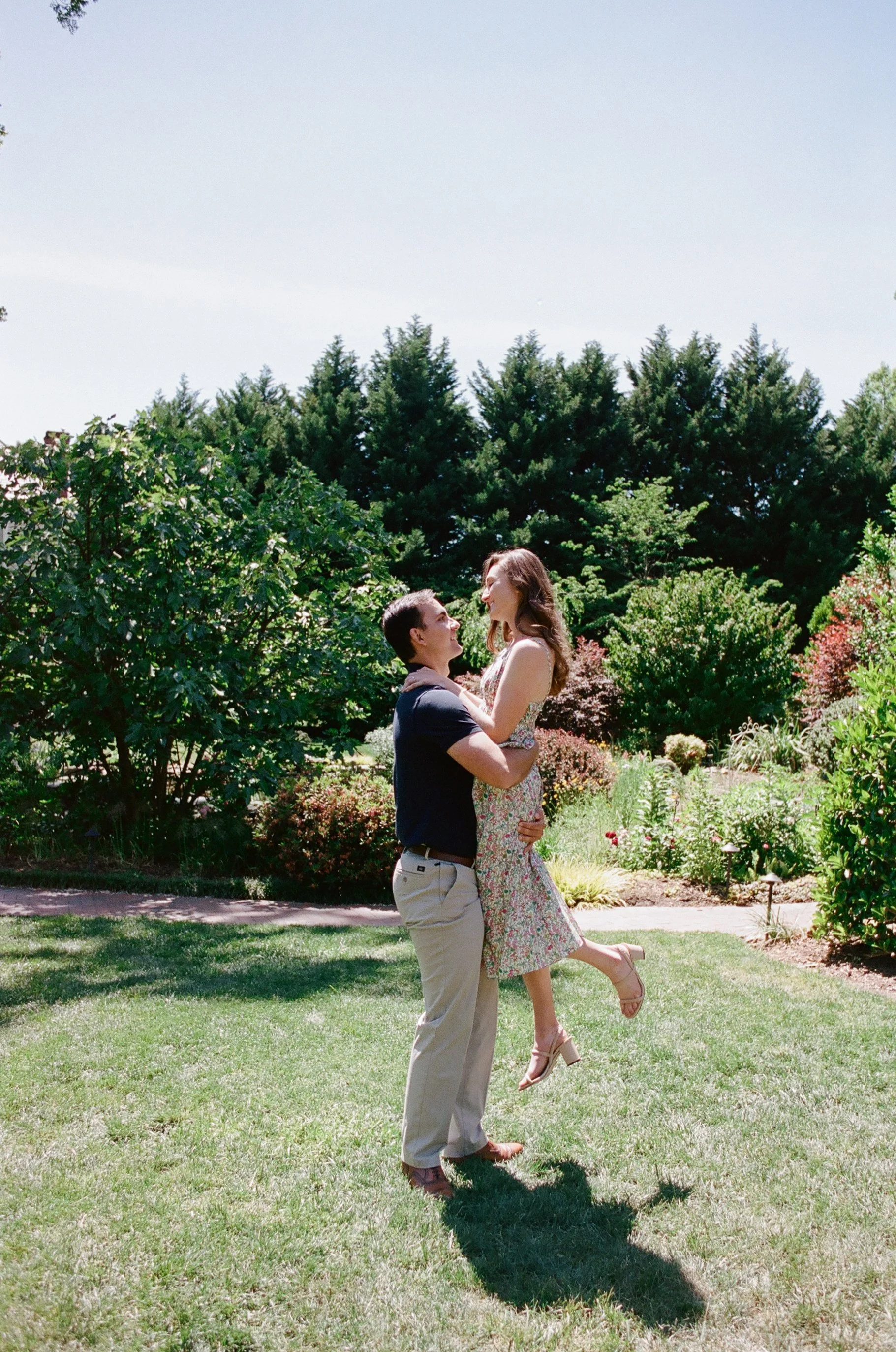 Guy swinging his fiancé in a summer dress in a field by private gardens at engagement session photographed on Kodak Portra 400 film at Alexander Homestead by Charlotte Wedding Photographer Michael Parks, owner of ParksPictures