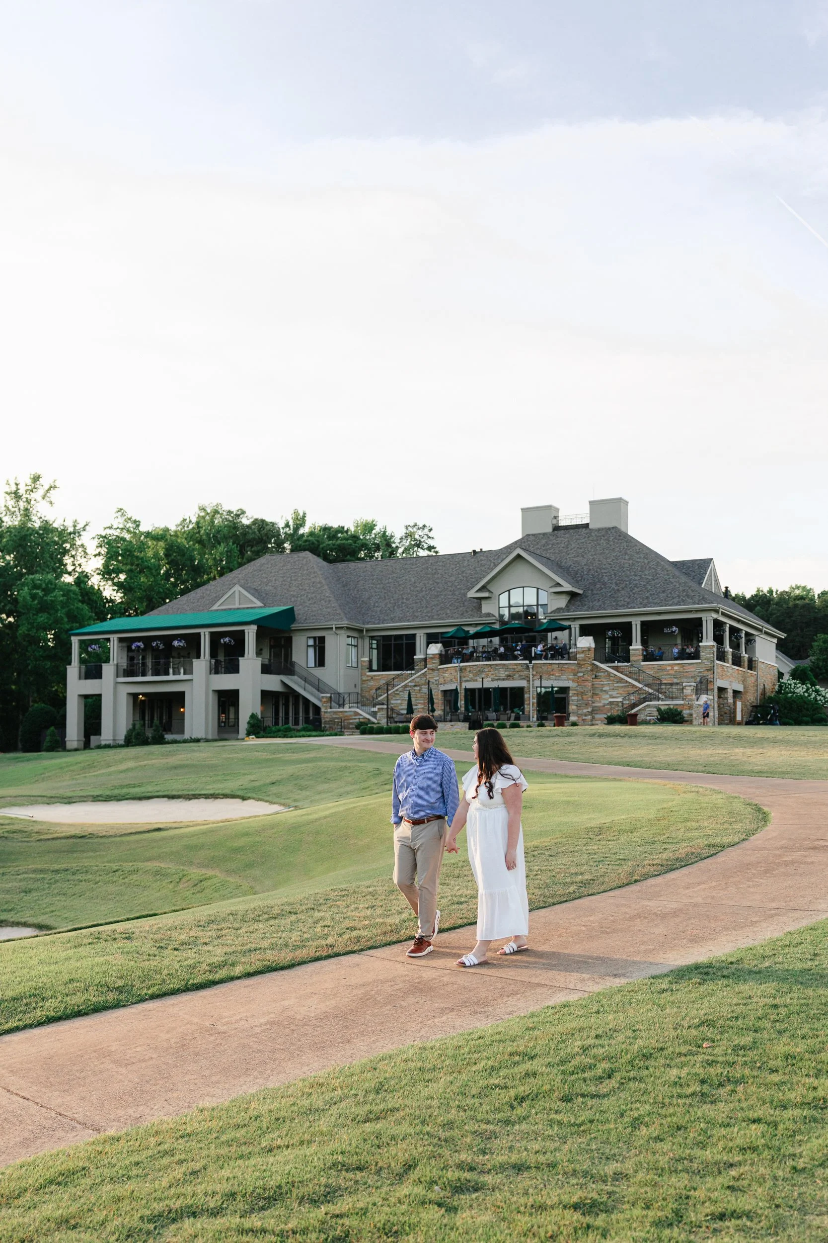 Guy and his fiancee holding hands and walking on golf course in front of clubhouse at NorthStone Country Club at sunset on film in a bright and airy aesthetic by ParksPictures
