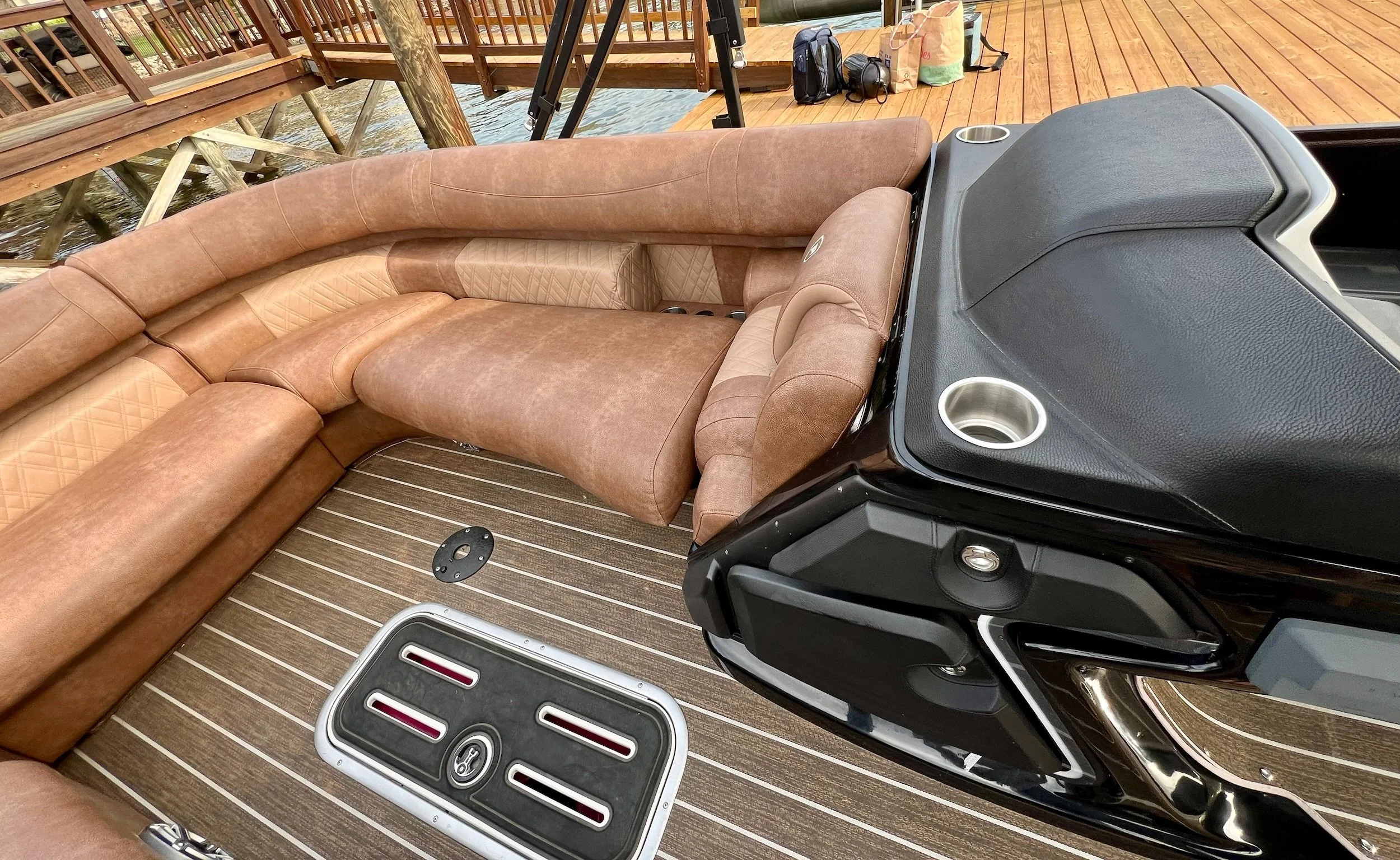 View of a boat's interior showing brown upholstered seating, a black dashboard with cup holders, and a brown and beige wooden floor, with a dock and water visible outside.