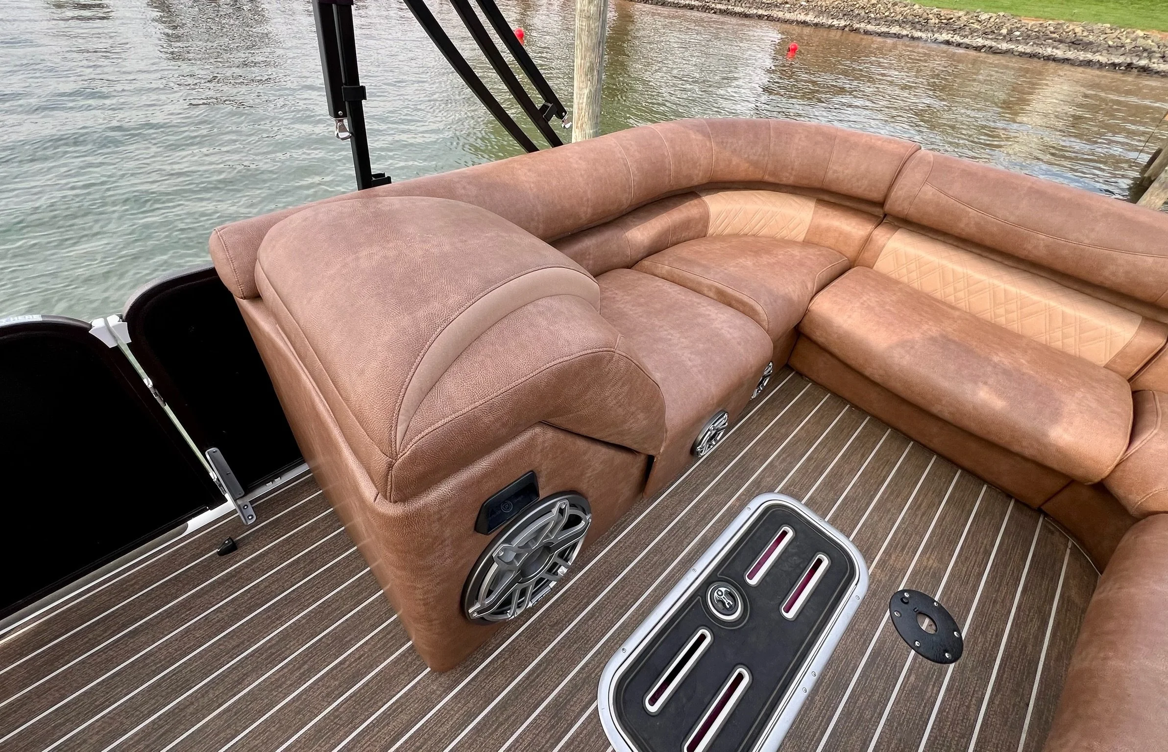 Brown cushioned seating area on a boat with speakers and a small black hatch in the deck, against water and a shoreline in the background.