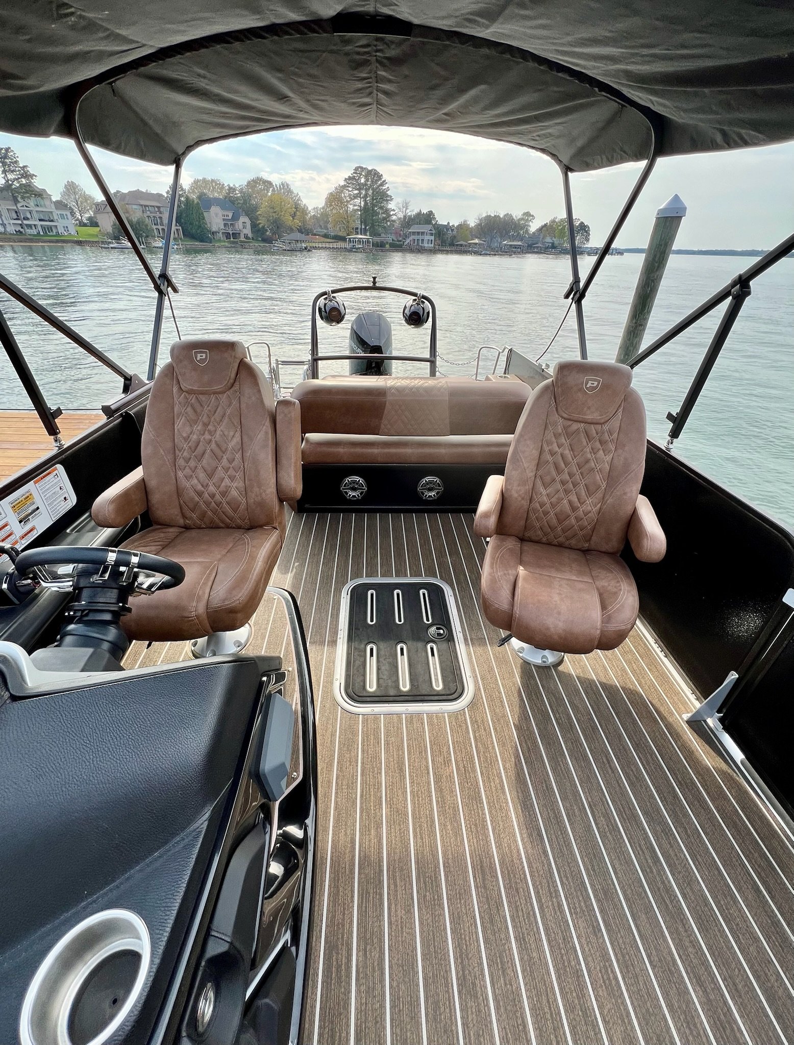 View of a boat's interior showing two brown cushioned captain's chairs, a steering wheel, and a dashboard with controls, with water and houses visible in the background.