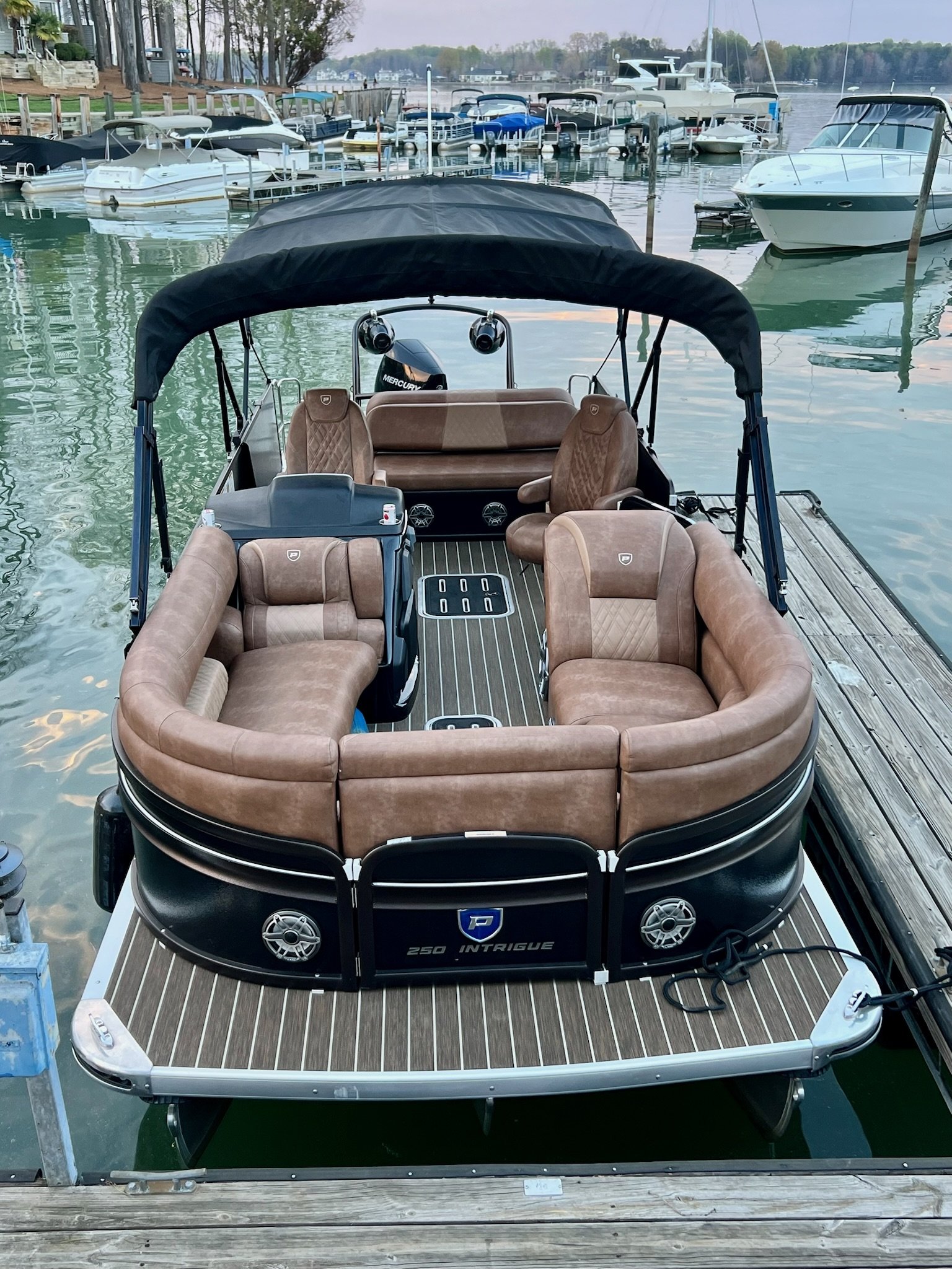 A pontoon boat docked at a marina, with brown leather seats arranged in a U-shape and a small steering area, surrounded by other boats and water.