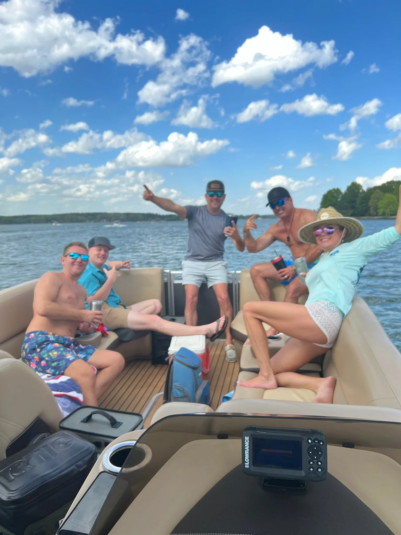 Group of six friends enjoying a day on a boat on the water, with a blue sky and fluffy clouds, some wearing swimwear and sunglasses, smiling and posing for the photo.