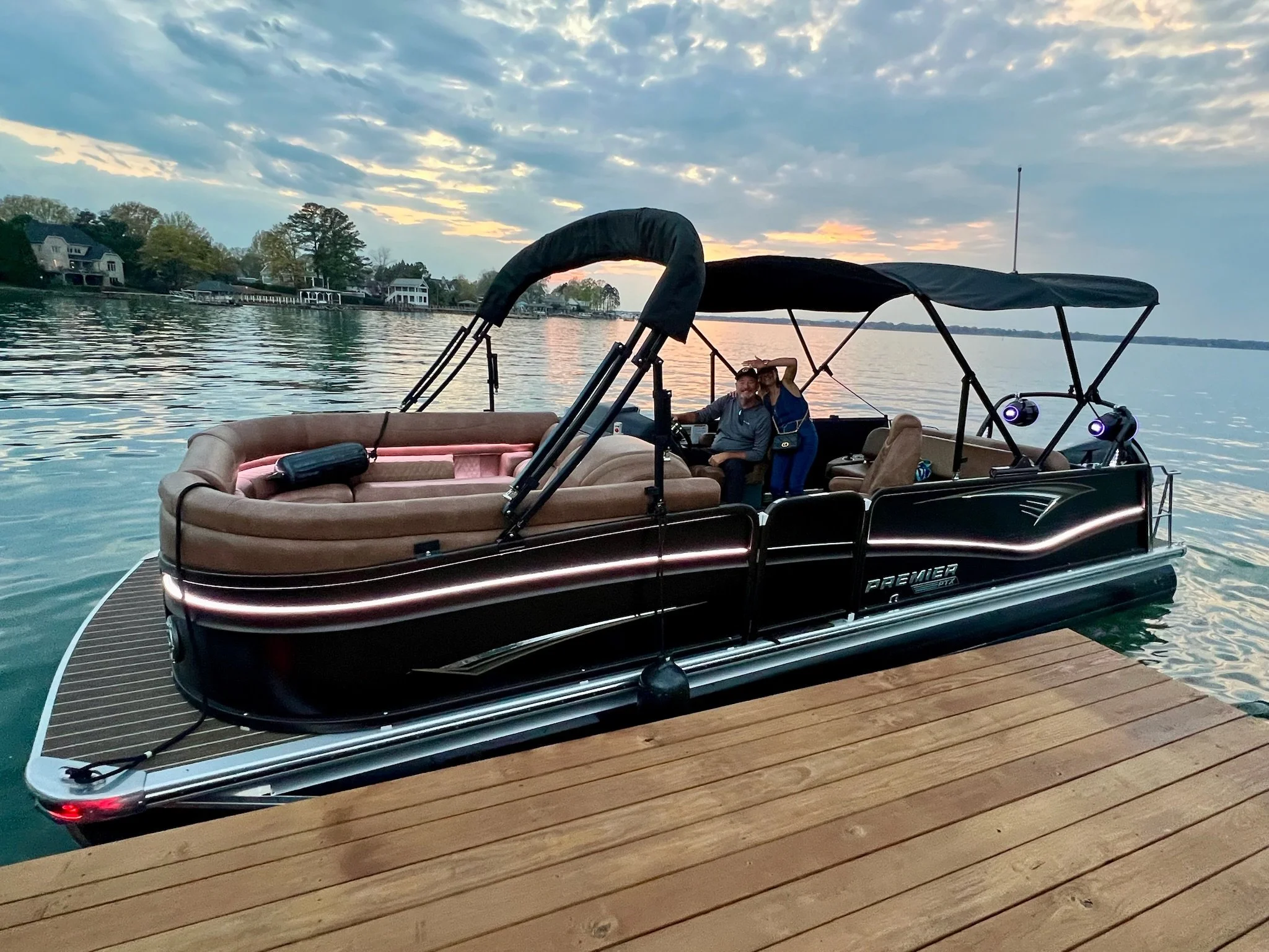 A black and brown pontoon boat docked at a wooden pier on a lake during sunset. There are two people inside the boat, smiling and waving. The background features a calm lake, houses along the shoreline, and a partly cloudy sky with the sun setting.