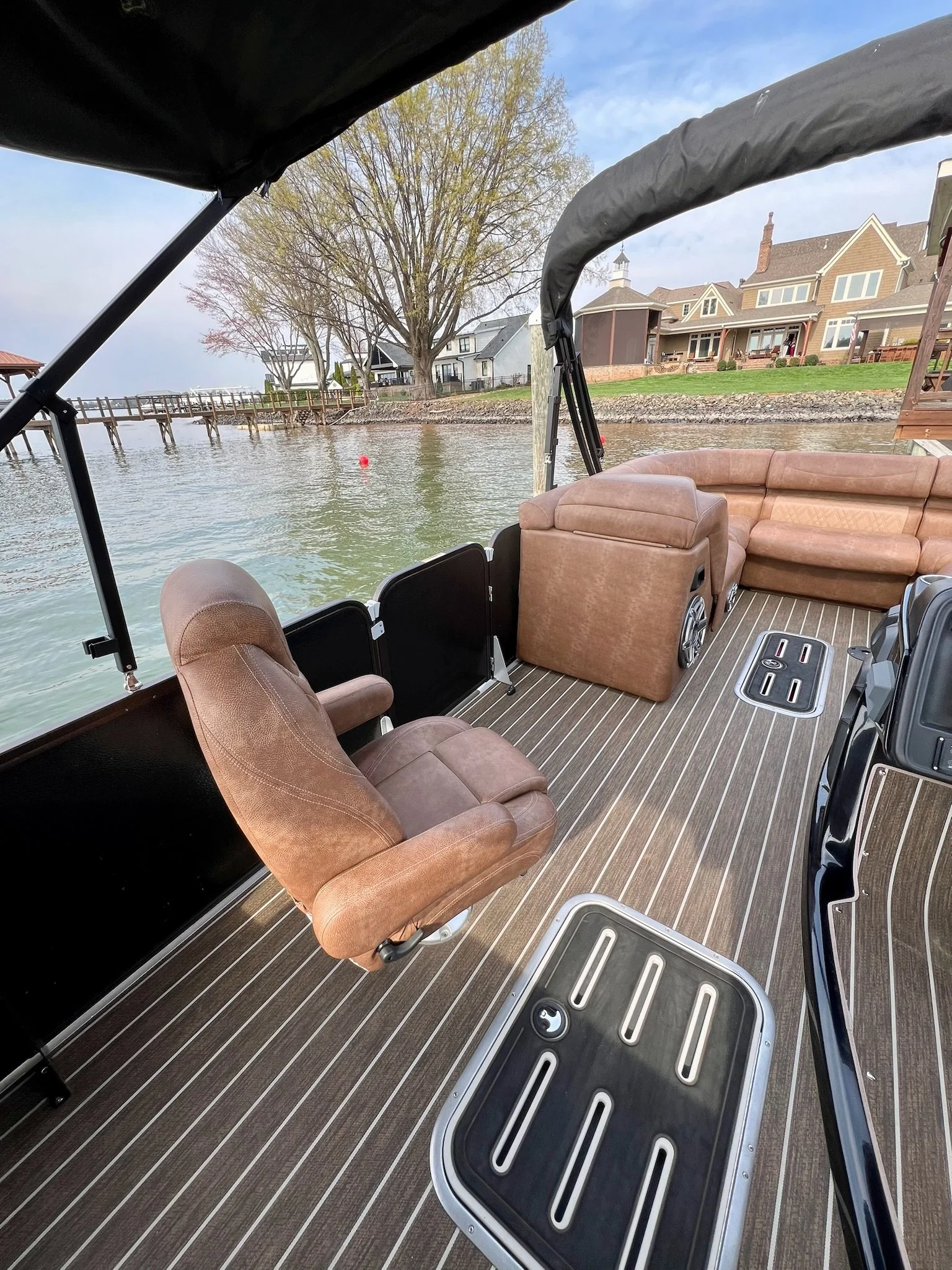 View of the deck of a houseboat, featuring brown upholstered seats, a table, and a scenic lakeside view with houses, trees, and a dock in the background.