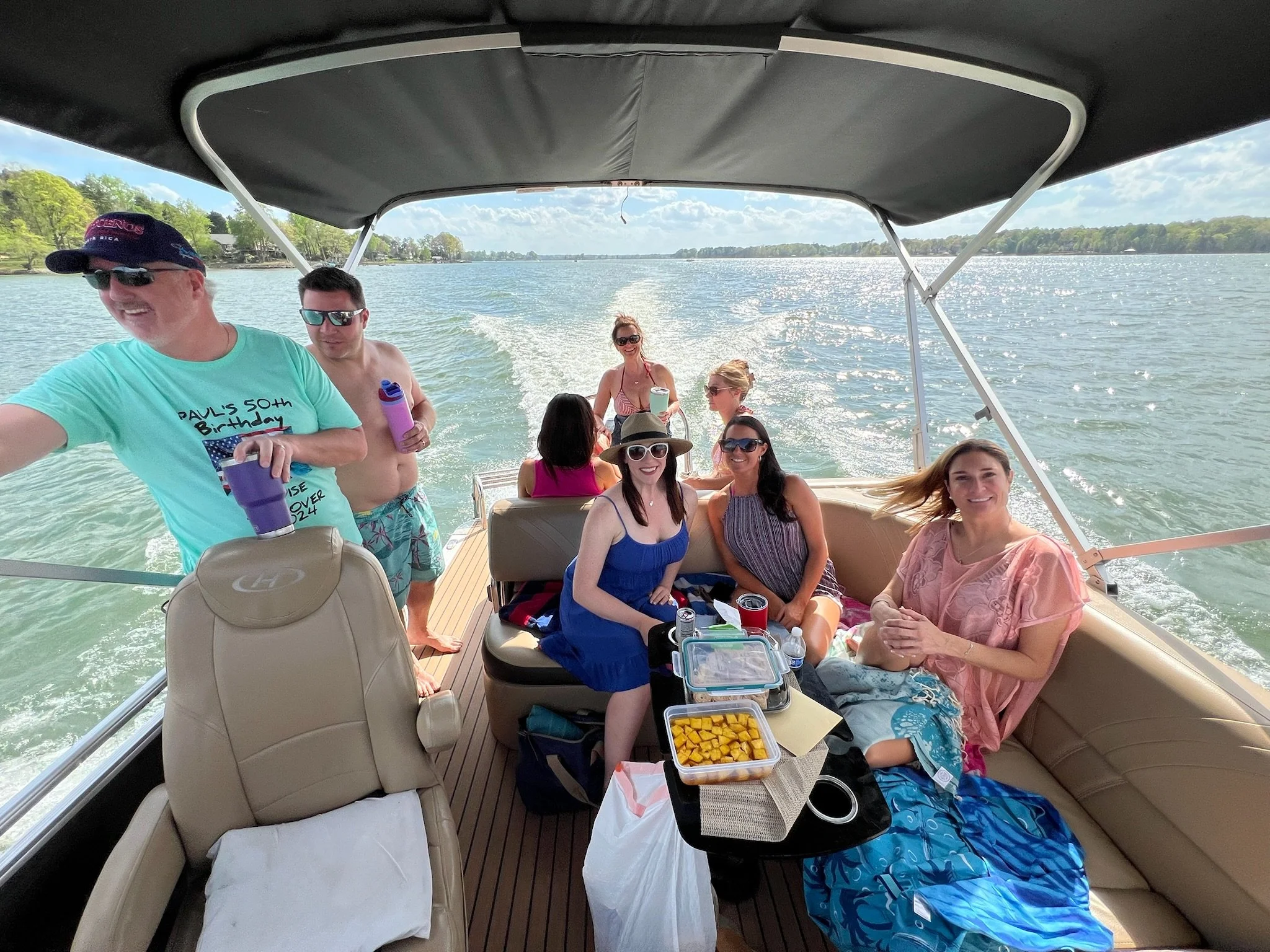 Group of people enjoying a boat ride on a lake, with some sitting on the boat's seats and others standing. The boat is moving through the water, creating a wake, and the sky is partly cloudy with a scenic view of trees along the shoreline.