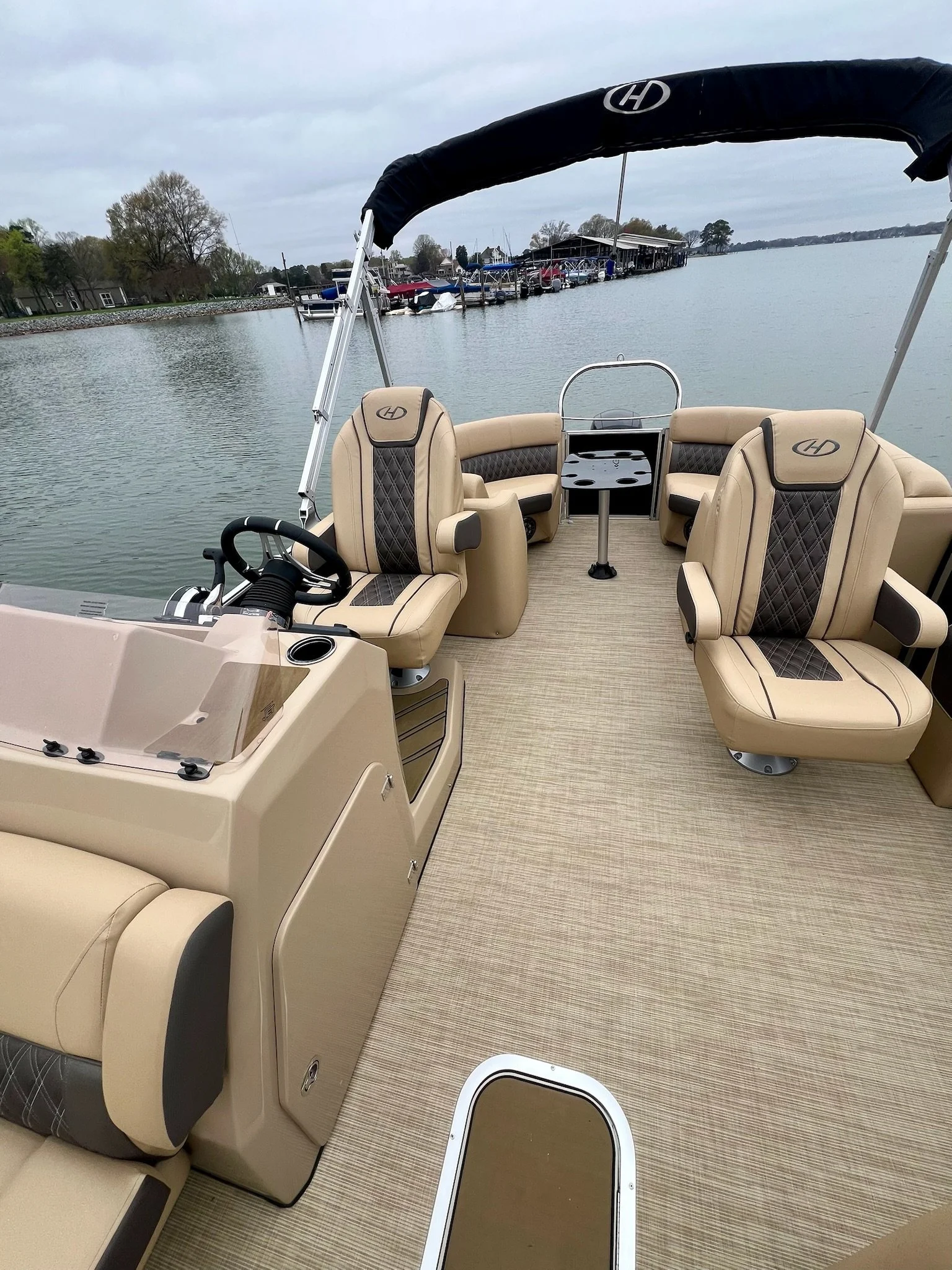 Captain's area of a beige pontoon boat with two captain chairs, a steering wheel, and a small table, on a calm lake with docked boats and a cloudy sky in the background.