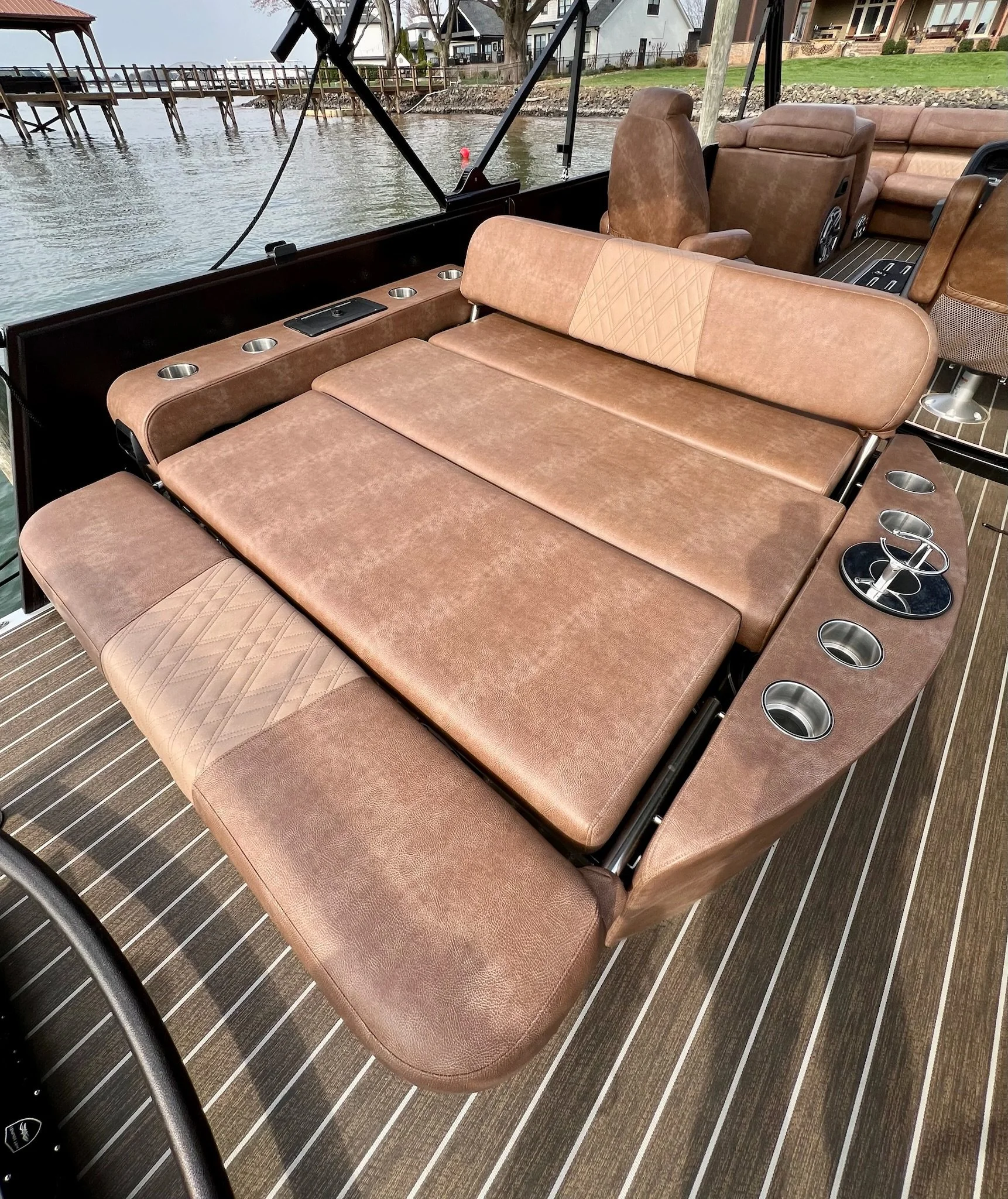 A boat deck with brown leather cushioned seating that can convert into a bed, cup holders, and scenic water and houses in the background.