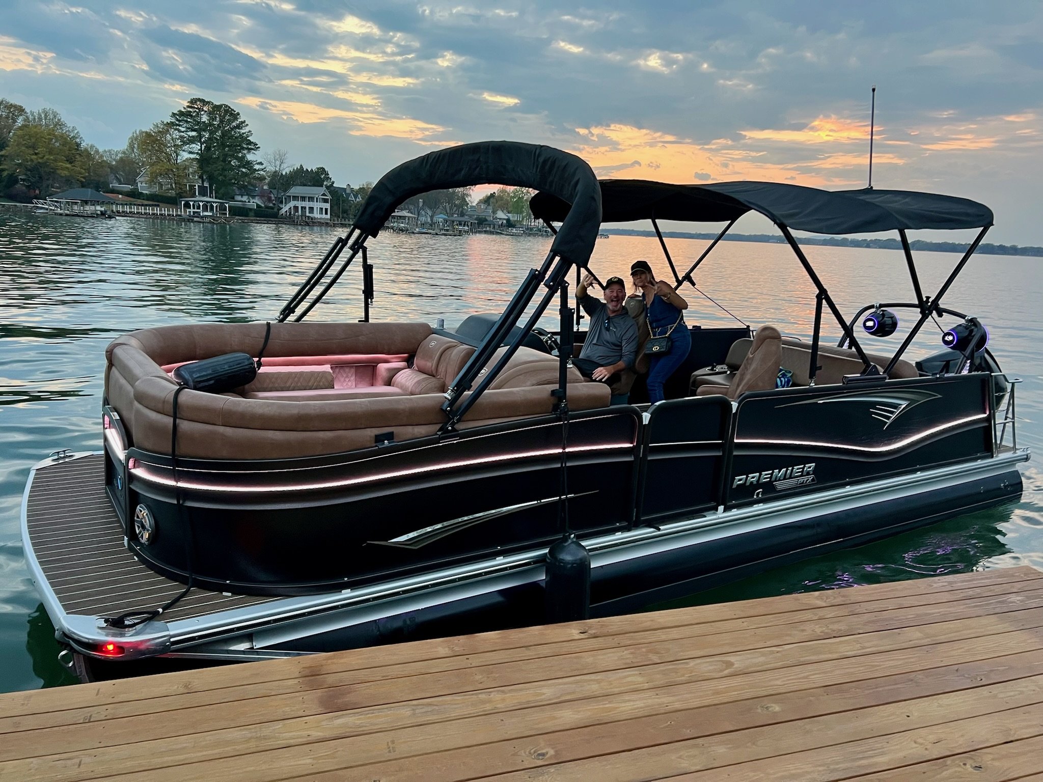 A pontoon boat floating on a lake at sunset with two people smiling and making peace signs, with houses and trees in the background.