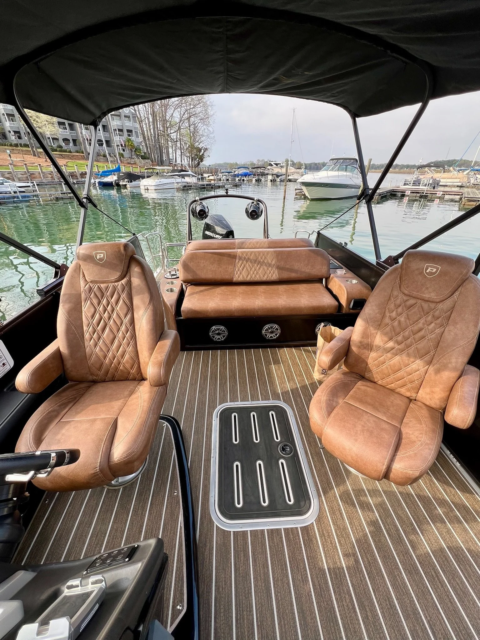Interior of a boat with two tan leather captain chairs, a brown leather bench seat, and a striped floor, overlooking a marina with multiple boats on the water.