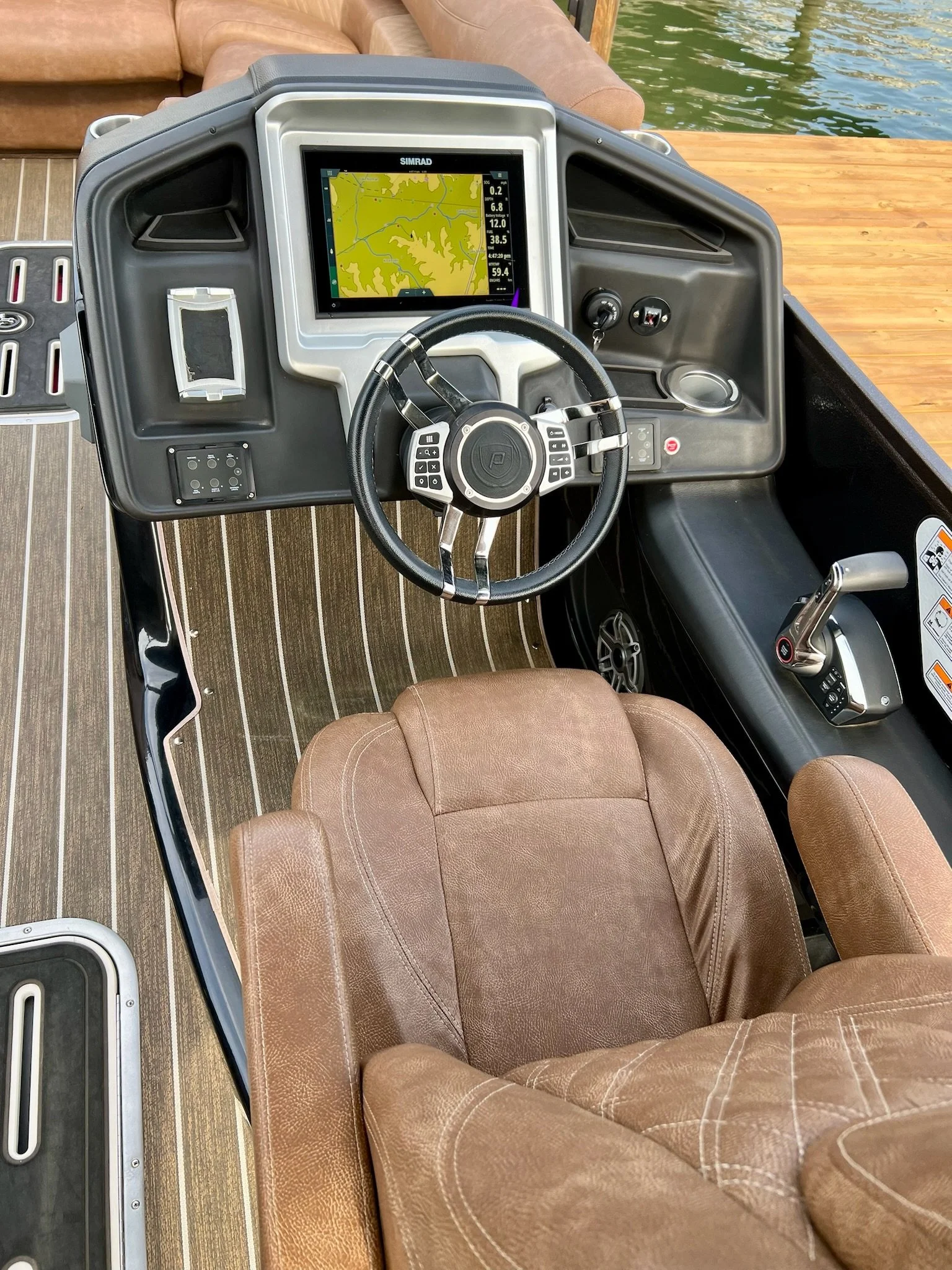 The image shows the cockpit of a boat with a steering wheel, a screen displaying a navigation map, and various controls. The boat is docked near a wooden pier, with water visible in the background.