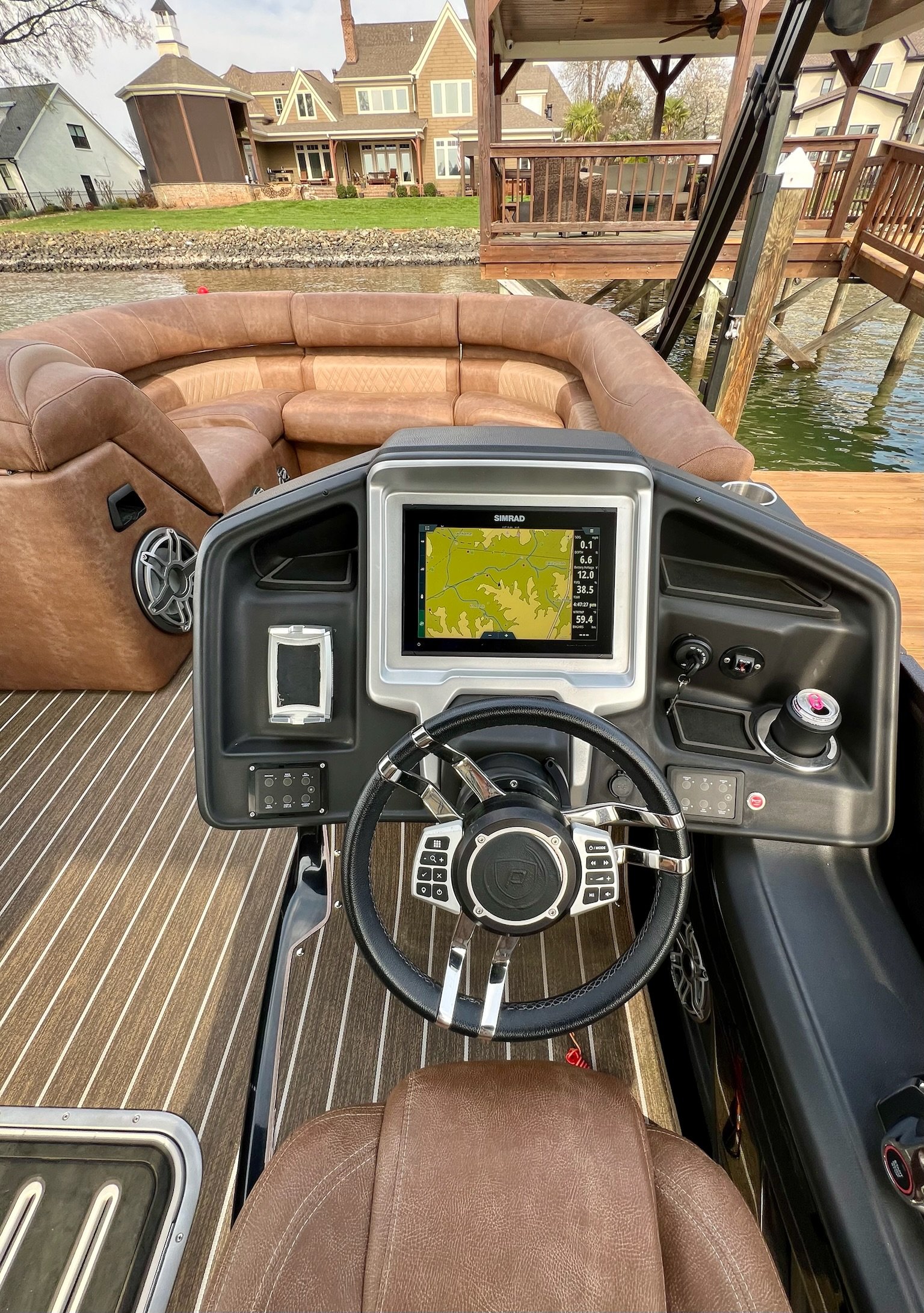The image depicts the helm of a boat with a brown leather captain's chair in front of a curved brown leather seating area. The dashboard features a GPS screen showing a map, along with various controls and a cup holder. In the background, a lakeside 