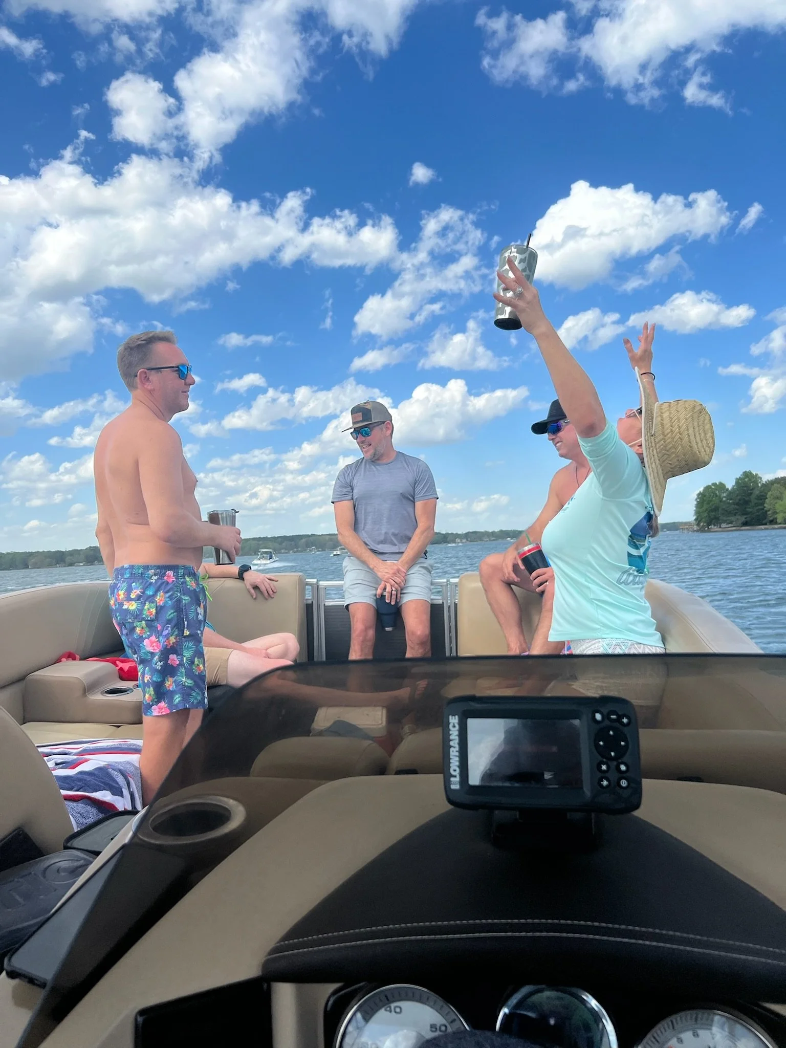 Group of friends on boat enjoying sunny day, some standing, some sitting, with drinks, water and sky in background