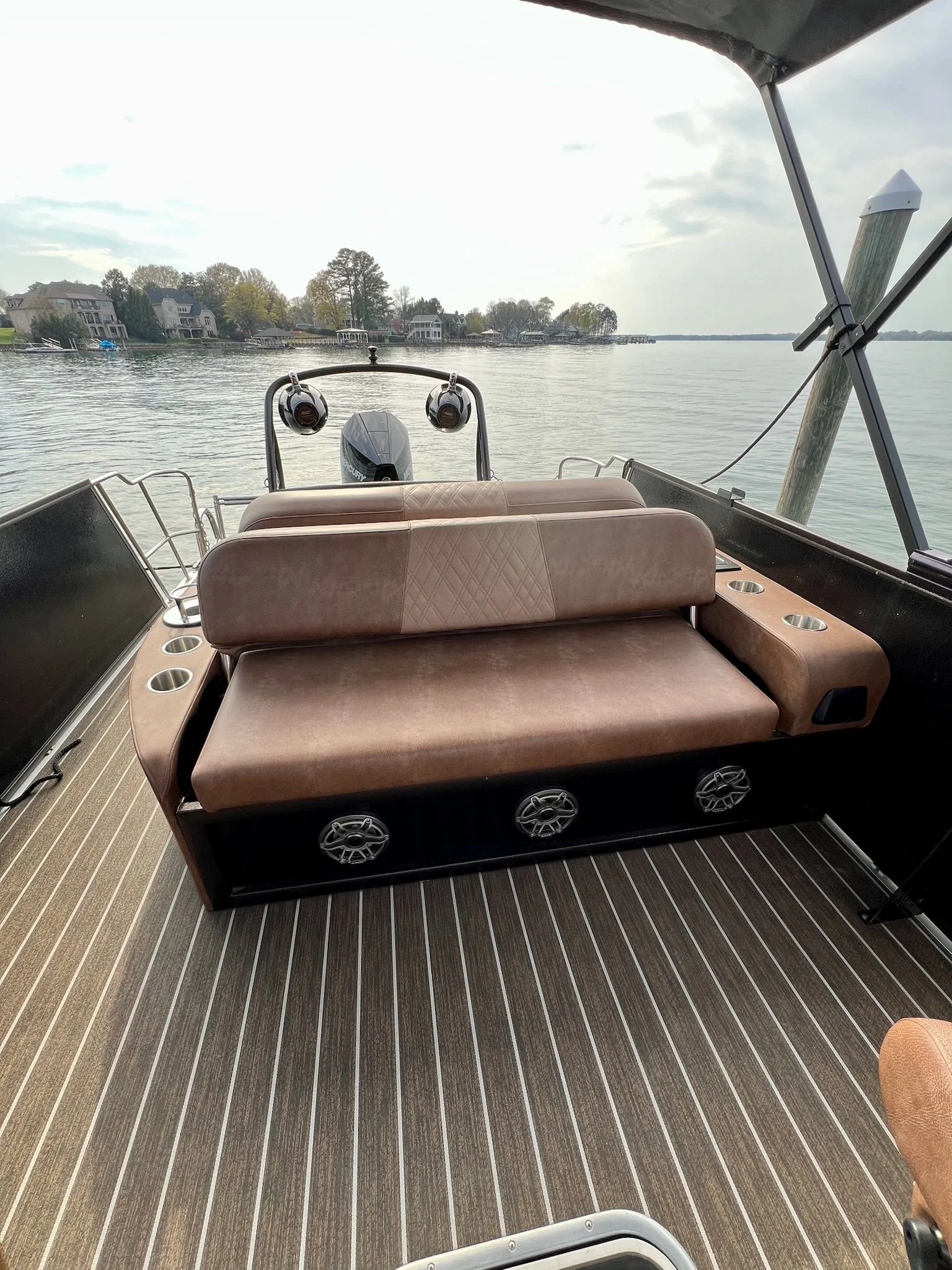 View of the outdoor living area on a boat, showing a brown cushioned bench, built-in cupholders, and a boat motor with water and houses in the background.