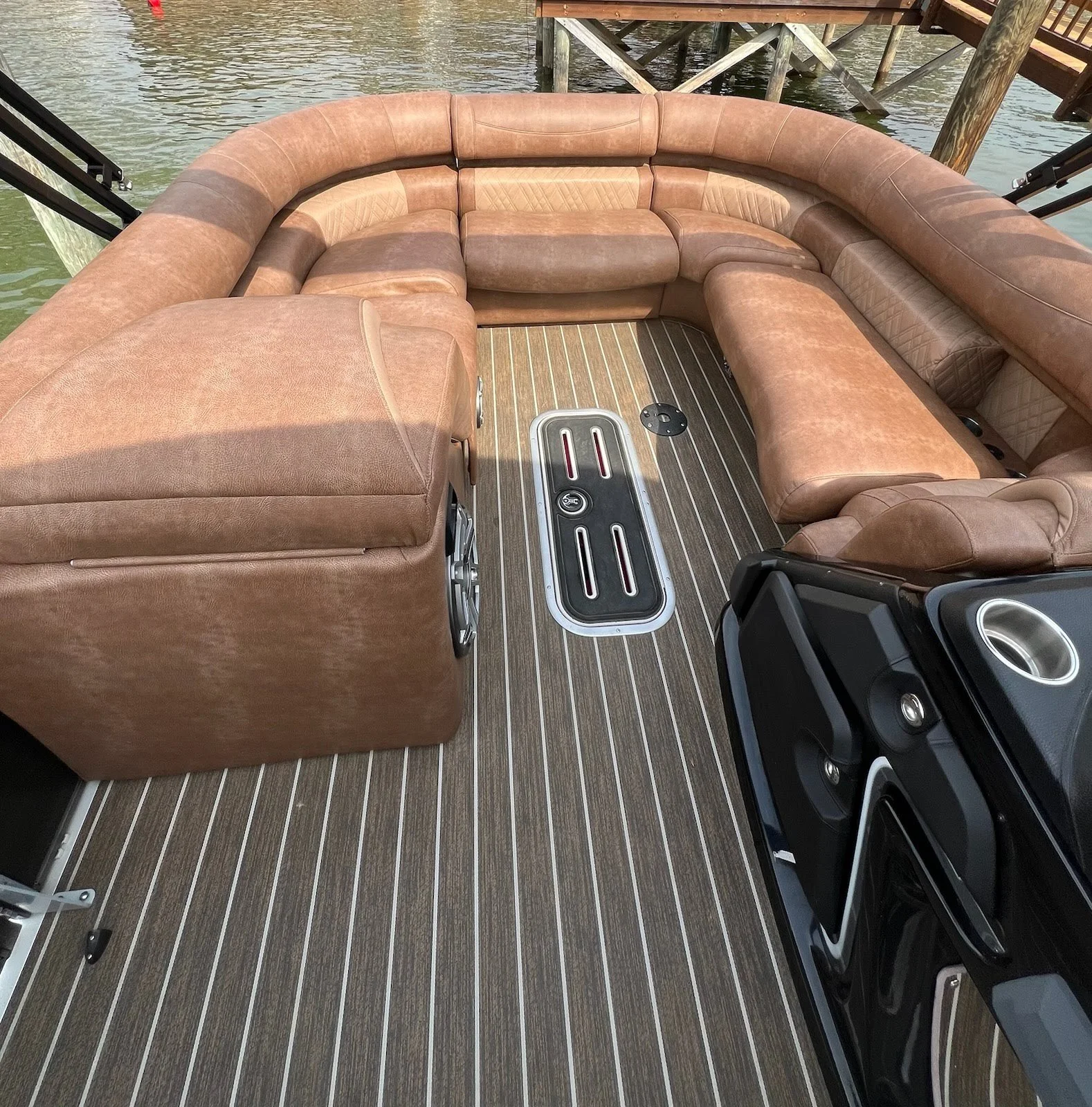 Interior of a pontoon boat with brown leather seating arranged in a U-shape and a wood laminate floor, near a dock with water visible in the background.