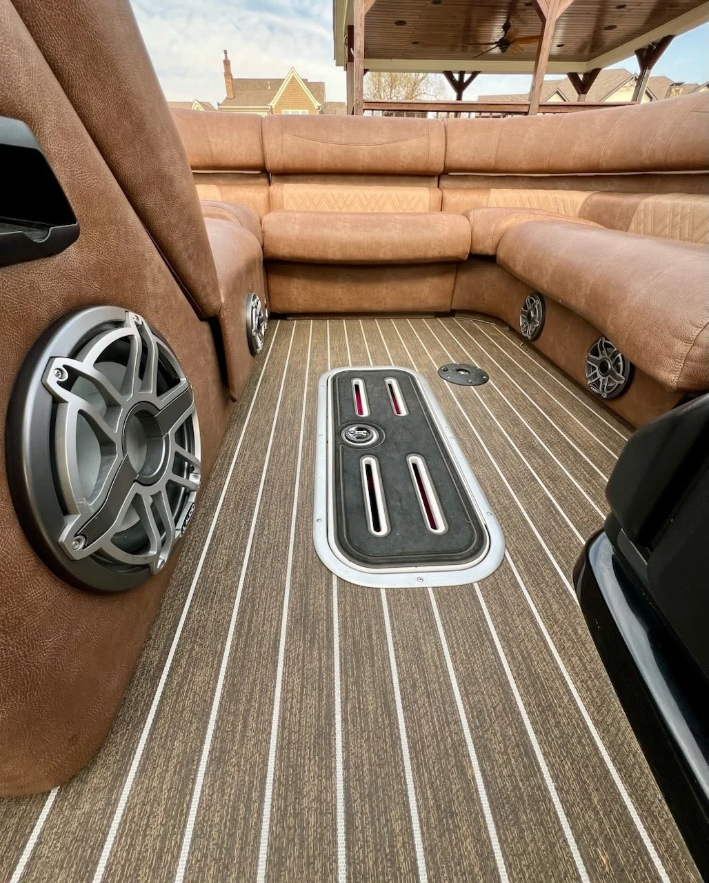 Inside a boat with brown leather upholstery, featuring large round speakers on the walls, a striped carpet floor, and a rectangular hatch in the floor, with views of houses and a porch outside.